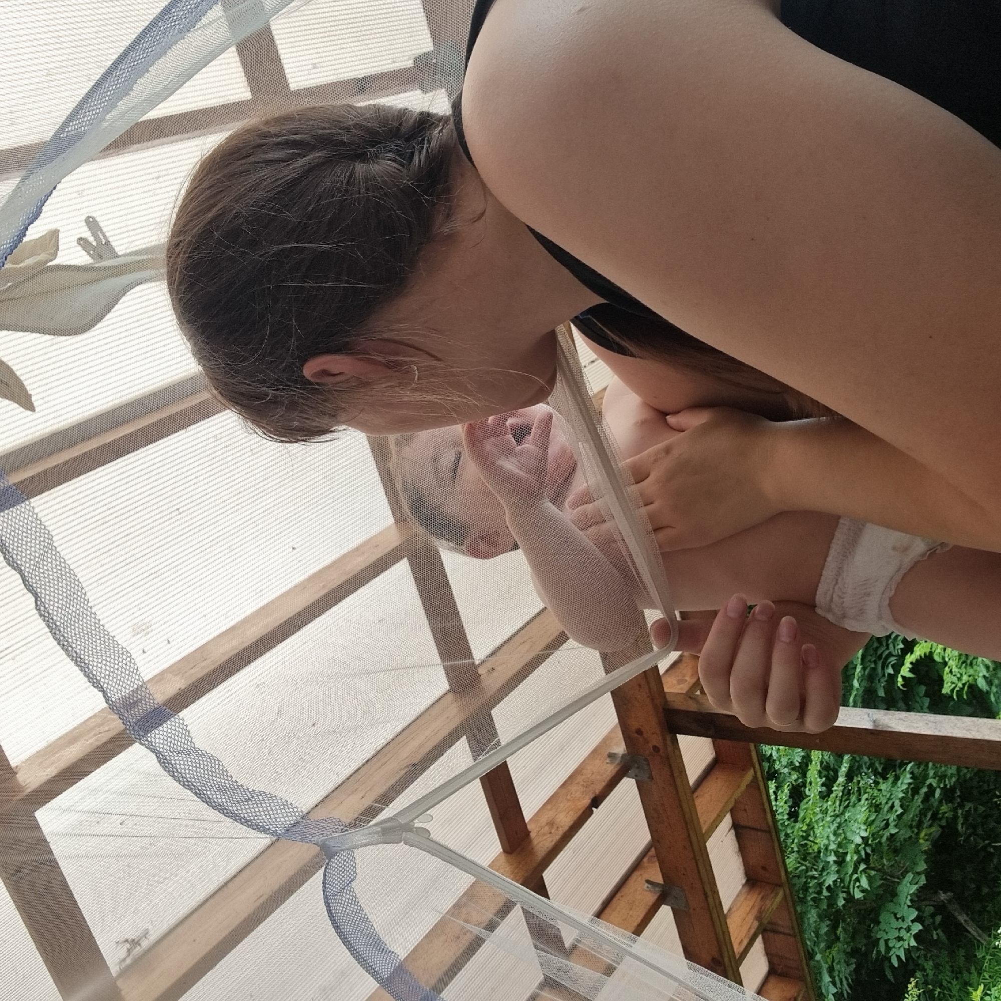 A young child, wearing a diaper, is held by an adult female under a netted canopy. The child's face is turned towards the adult, with their mouth open as if crying. The adult's face is partially visible, with their head tilted down towards the child. The canopy is suspended from wooden beams and a translucent roofing material. Lush green foliage is visible in the background, suggesting an outdoor setting. The scene is captured from a low angle, looking upwards. There is no information in the image to suggest the location is Dunnae-myeon, South Korea.