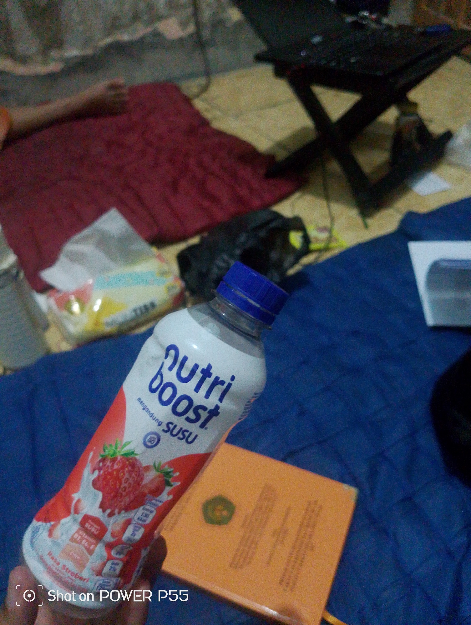 In a dimly lit indoor environment, a person's hand holds a white bottle of Nutriboost strawberry milk. The bottle features a blue cap and a label with images of strawberries and nutritional information. The background includes a red cloth, a black object that appears to be a laptop stand with a laptop on it, and a dark blue surface that might be a mat or blanket. An orange book with text is also visible. The timestamp "Shot on POWER P55" is present in the bottom left corner. The location is identified as Lebaksiu, Indonesia.