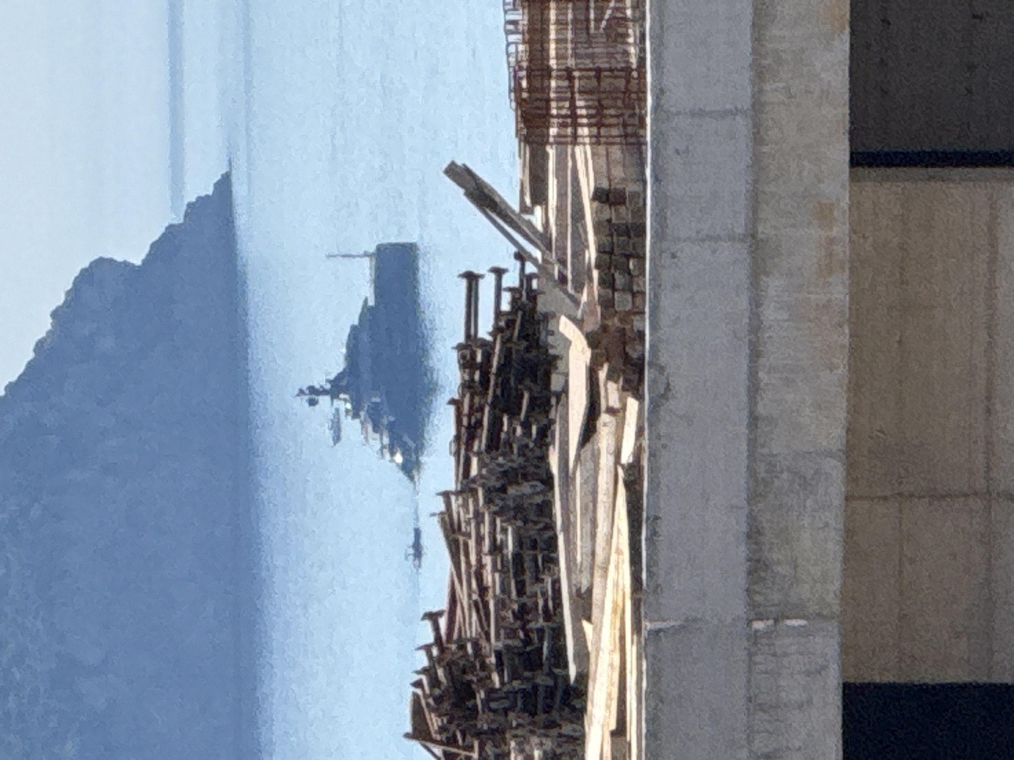 In Rafailovići, Montenegro, a large, dark-colored superyacht is visible anchored in the clear blue sea. In the foreground, a construction site is apparent, featuring stacks of wooden planks and what appears to be rebar reinforcement. A rugged, tree-covered hillside dominates the background, with the sea extending to the horizon. The overall scene depicts a juxtaposition of leisure and development along the Montenegrin coastline.