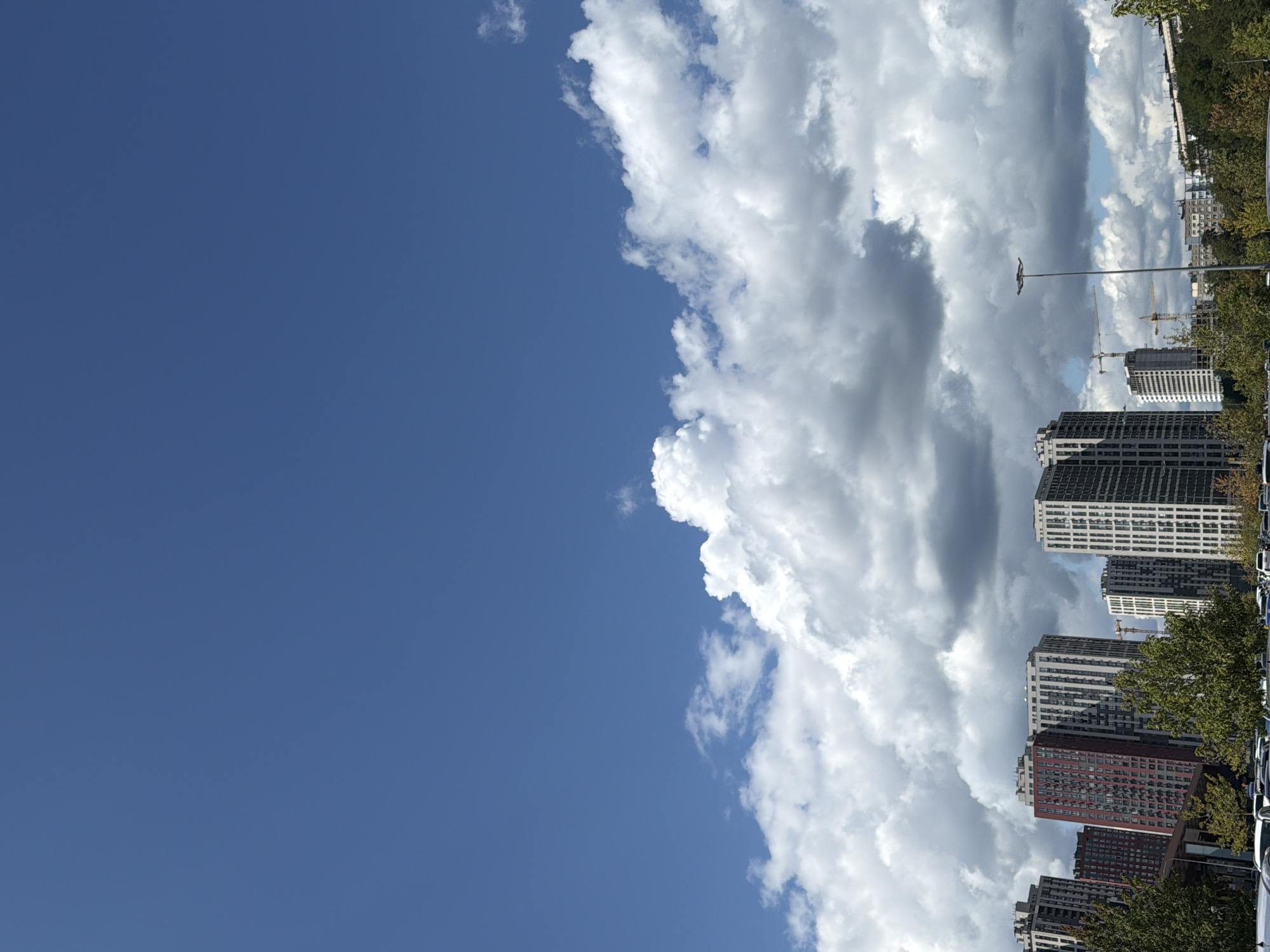 A wide shot captures a cityscape with multiple modern high-rise buildings under a vast, bright blue sky with scattered white cumulus clouds. In the foreground, a parking lot is partially visible with several cars. The buildings, predominantly residential, vary in height and architectural style, with some featuring red and white facades, while others are clad in gray and white panels. Green trees are interspersed among the buildings and in the foreground, suggesting an urban park or residential area. The scene depicts a typical day in an urban environment, possibly in Kyiv, Ukraine, given the architectural styles and common urban features.