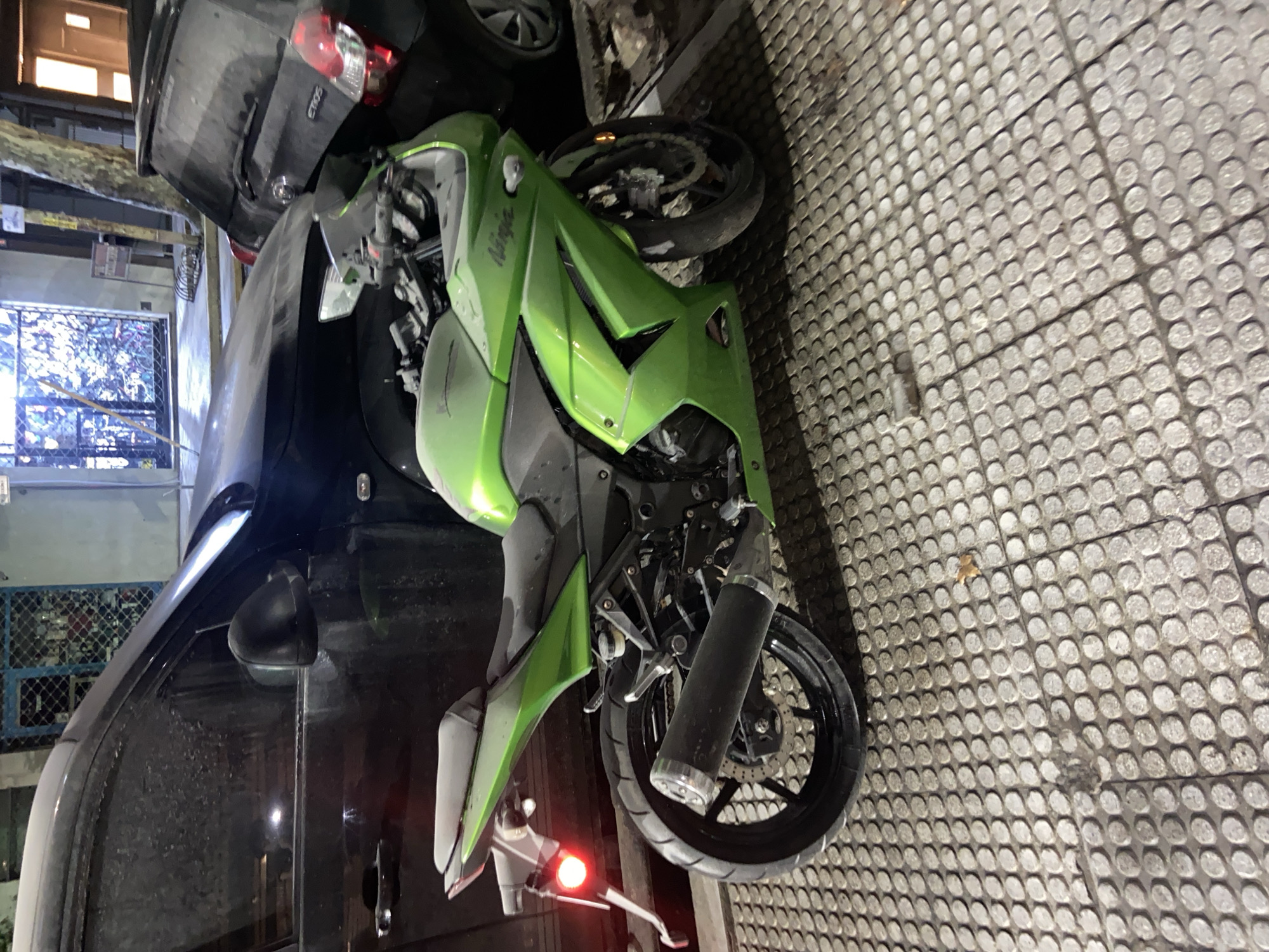 A green Kawasaki Ninja motorcycle lies on its side on a paved walkway in what appears to be an urban environment. The motorcycle is entangled with a dark-colored vehicle, possibly a car, positioned vertically. The paving stones on the walkway have a textured, dotted pattern. In the background, there are buildings, a window with a metal grill, and what seems to be tree foliage. The location is suggested to be in Buenos Aires, Argentina, though there are no obvious location cues in the image. The scene appears to depict the aftermath of an accident or an unusual parking situation. No people are visible in the image.