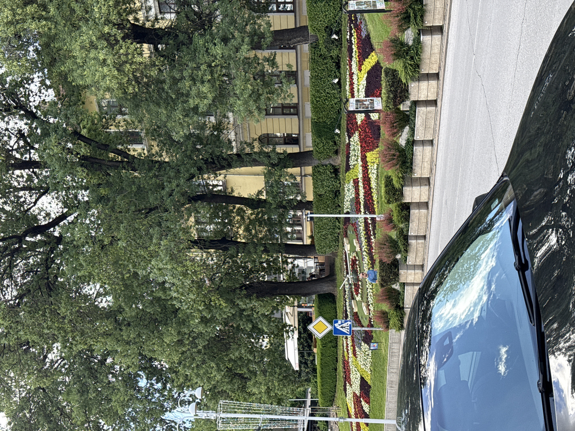 The image, taken from inside a dark-colored car, depicts a street scene in Veliko Tarnovo, Bulgaria. Through the car's windshield, one can see a meticulously arranged flower bed along the sidewalk. This flower bed features a colorful display of red, white, and yellow flowers. A hedge serves as a backdrop to the flower bed, with trees providing further shade.
Road signs are visible along the street, indicating traffic regulations or directions. Beyond the trees, a light-yellow building with dark-framed windows can be seen. A figure of a person is standing near the flowerbed. A decorative lamppost is visible in the foreground.