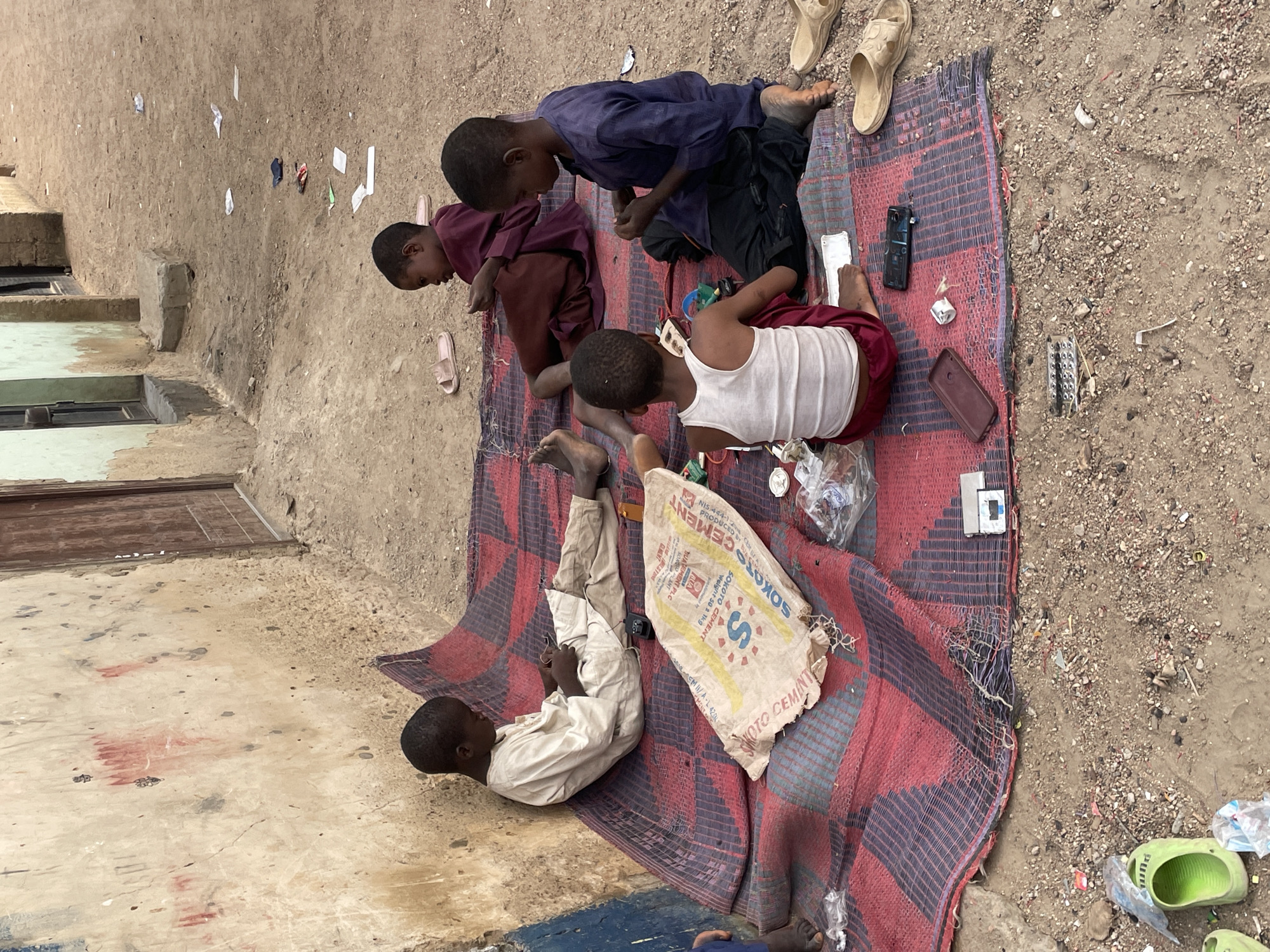 The image shows a group of children playing on a weathered, red and purple striped mat laid out on a dirt surface. One child is lying on their back, seemingly engrossed in a mobile phone. Another child beside them is close to a torn sack labeled "SOKOTO CEMENT." A cluster of three more children are kneeling and focused on something on the mat. Discarded items such as shoes, bits of plastic, a pair of sandals, and various metallic and plastic objects surround the mat. The backdrop reveals an old building with weathered walls and windows.