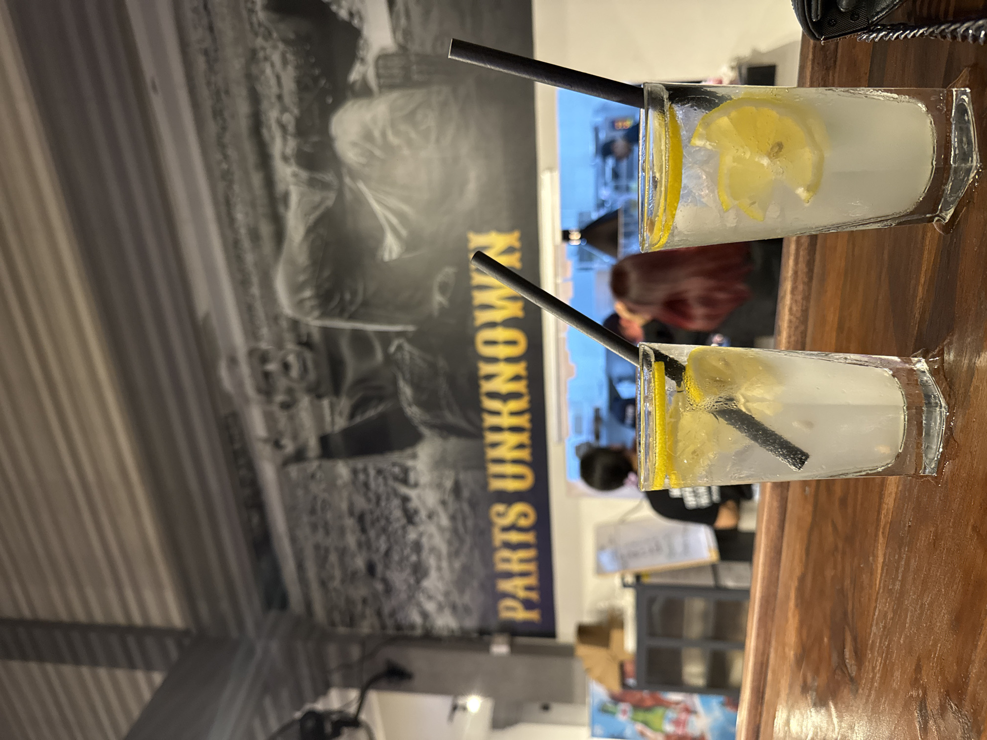 Two glasses of lemonade with ice and lemon slices and black straws sit on a wooden bar counter. In the background, a large black and white mural depicting figures is displayed on the wall above text that reads "PARTS UNKNOWN". The environment appears to be an indoor establishment, possibly a bar or restaurant, with other patrons visible in the background. The lighting is dim, creating a relaxed atmosphere.