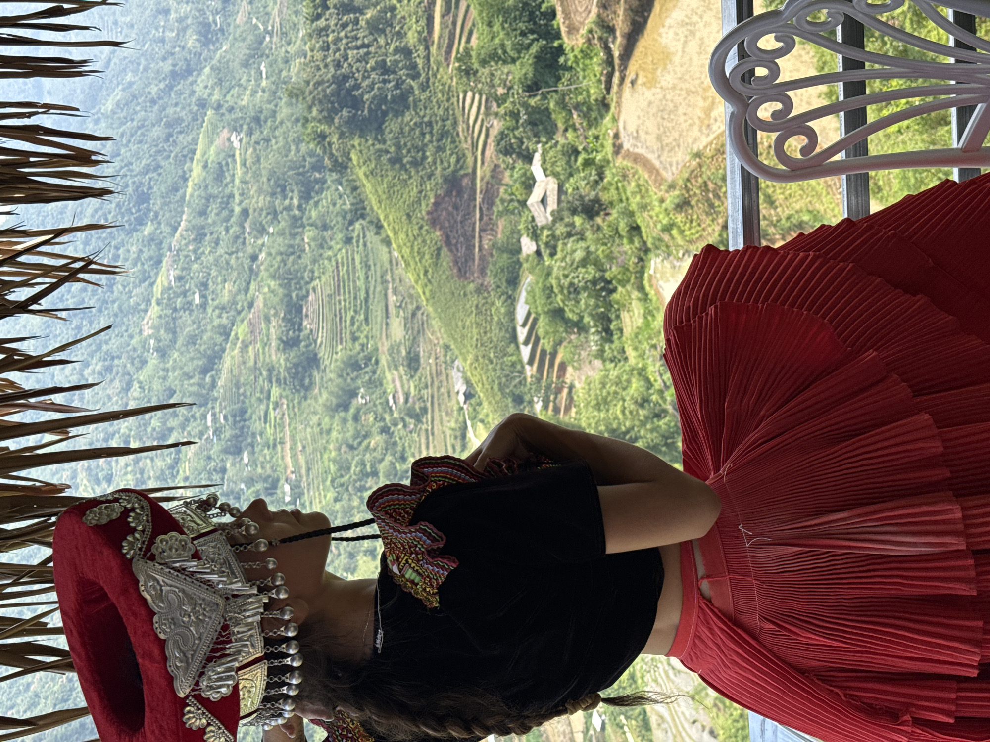 A woman in traditional attire is captured in a high-angle shot, looking up and out over a vast mountainous landscape in Thông Nguyên Commune, Vietnam. She is adorned with a prominent red velvet hat featuring elaborate silver metalwork and dangling silver bead ornaments. She wears a black top with colorful embroidered details on the shoulders and a voluminous, pleated red skirt. Her dark hair appears to be braided or styled with strands falling forward.
In the immediate foreground to the left, natural elements resembling dried straw or leaves from a thatched structure are visible. To the right, part of a white, ornate metal chair back suggests she is positioned in an outdoor seating area or balcony overlooking the scene.
The background is dominated by lush green mountains carved with extensive rice terraces, some of which appear to be filled with water, creating reflective surfaces. Small buildings are scattered across the distant hillsides, hinting at human presence within the expansive natural setting. The lighting indicates a bright, clear day. The woman's posture suggests she is admiring the breathtaking panoramic view. No text is visible in the image.