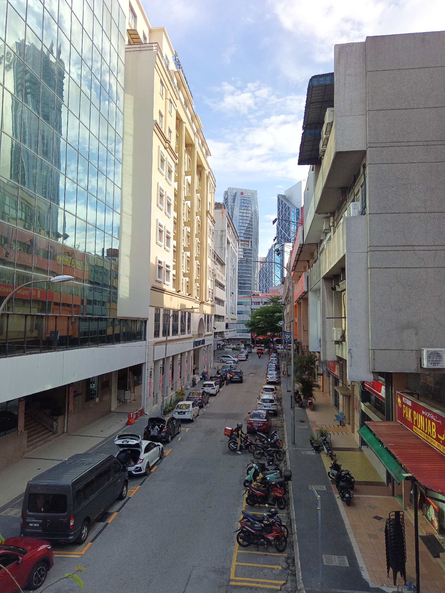 The image captures a street scene in Kuala Lumpur, Malaysia, likely in a commercial or mixed-use district. The perspective is from an elevated position, looking down onto a two-lane street lined with buildings on both sides.
On the left, a modern building with a facade of reflective glass windows dominates the foreground. The reflections show tall buildings, indicating a city environment. Adjacent to this is a lighter-colored, multi-story building with a more traditional architectural style, featuring repetitive window patterns and decorative elements.
The street itself is occupied by a line of parked vehicles, including several cars, a dark-colored van, and numerous motorcycles. Some cars have their trunks open. A few people are visible on the sidewalks.
On the right side of the street, the buildings appear older, with concrete facades and awnings over storefronts. One visible storefront has signage for "PAK PUNJAB RESTORAN". The sidewalk on this side is paved with tiles.
The sky above is partly cloudy with a bright, overcast light, suggesting it is daytime. The overall impression is of a busy urban environment with a mix of modern and older architecture. The presence of the van with license plate "VAW 3889" might be a clue to a specific location or vehicle registration details.