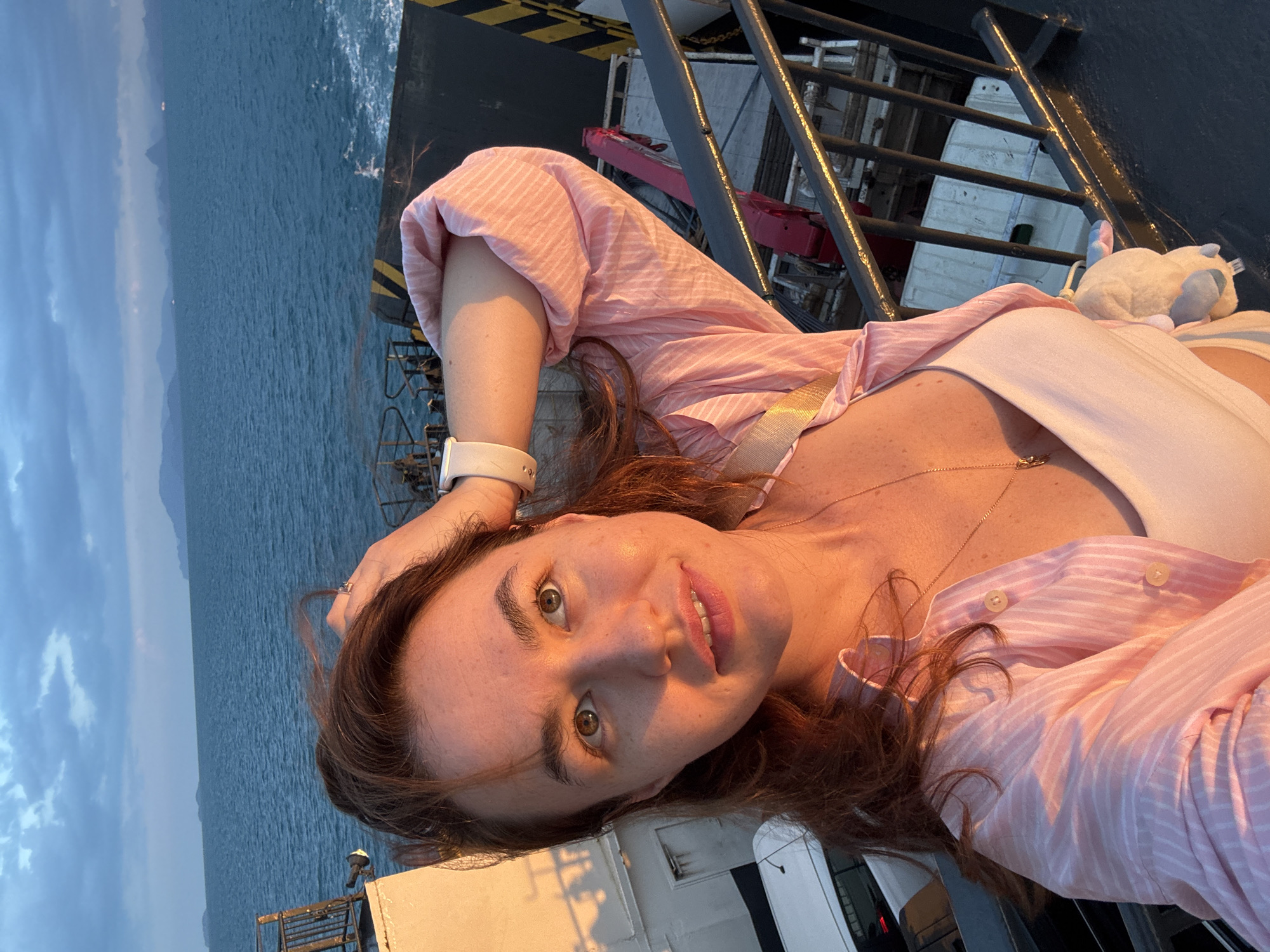 A woman with light brown hair is taking a selfie on the deck of a ferry. She is wearing a pink and white striped button-down shirt over a white tank top and a gold necklace. Her right arm is raised, with her hand resting on her head. The ferry is at sea, with calm blue water and a cloudy sky. In the distance, there are islands. The deck of the ferry is visible in the foreground, with vehicles and railing. The lighting suggests it is either sunrise or sunset. The location is identified as Unknown, Thailand.