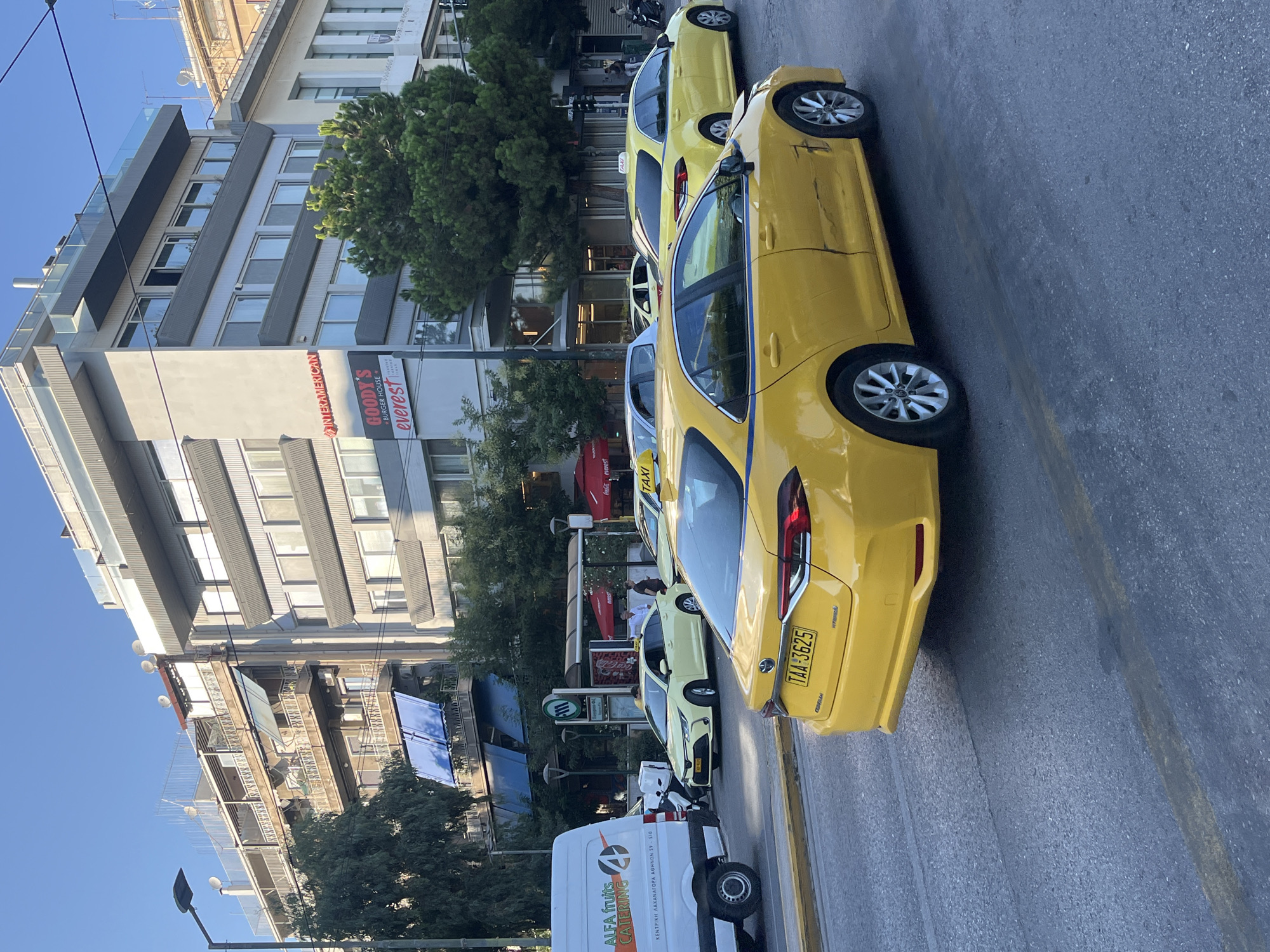 In Athens, Greece, a yellow taxi, identified by its license plate "TAA-3625", is the central focus of the image, situated on a city street. The taxi is a sedan and appears to have sustained damage to its rear passenger side. In the background, several other yellow taxis are visible, one with a "TAXI" sign on its roof, and a white van with the logo "ALFA fruits CATERING". The scene is framed by buildings, including a multi-story structure with balconies and awnings, and another building with signage that includes "INTERAMERICAN" and "GOODY'S BURGER HOUSE everest". Trees line the street, and overhead, power lines are visible. The sky is clear and blue, suggesting daytime.