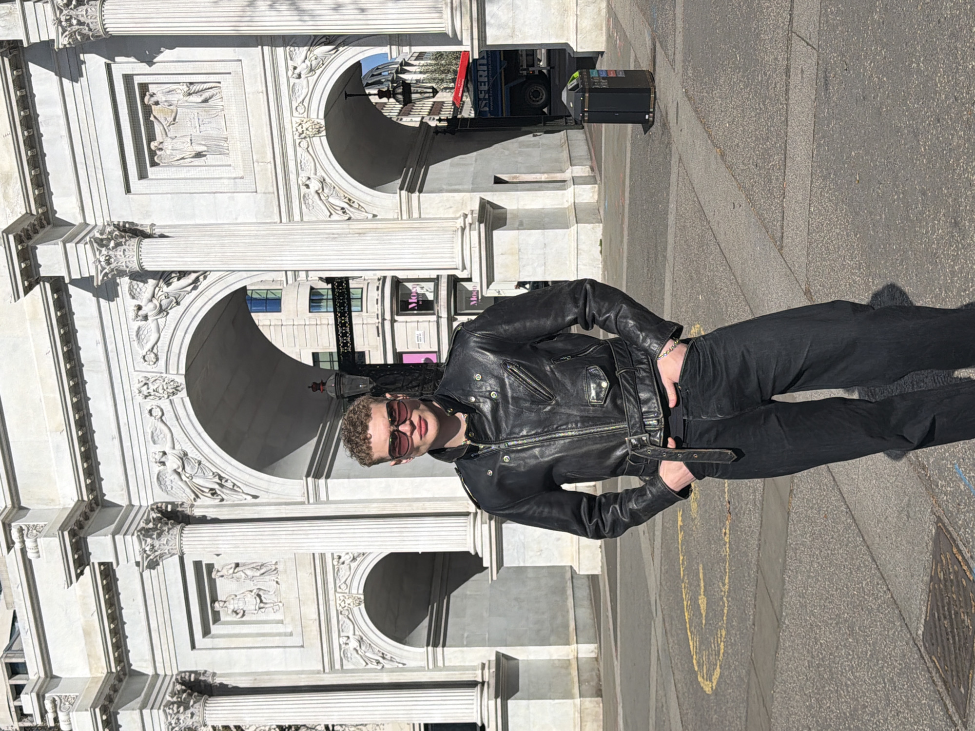 A young adult male stands in the foreground, centrally positioned, against the backdrop of the Marble Arch in London, United Kingdom. He is observed wearing dark sunglasses, a black leather motorcycle-style jacket with multiple zippers and a belt, and dark jeans. His hands are placed within his pockets, and his gaze is directed towards the camera. He has short, curly brown hair.
The Marble Arch, a large white marble triumphal arch, features three archways, fluted Corinthian columns, and various classical relief sculptures including winged figures holding wreaths and rectangular panels depicting draped individuals. The structure exhibits signs of weathering. Through the right archway, a partially visible blue truck with "ESSENTIAL" branding and a red roof element is present. A dark public litter bin is positioned to the right of the male subject, adjacent to the arch base. An ornate, dark lamppost hangs within the right archway. In the background, beyond the arch, modern urban buildings are visible, one displaying a "Moco" sign. The ground surface is paved with asphalt and lighter concrete sections, showing faded yellow road markings and a metal manhole cover on the left. The scene is illuminated by strong, direct sunlight, casting distinct shadows.
