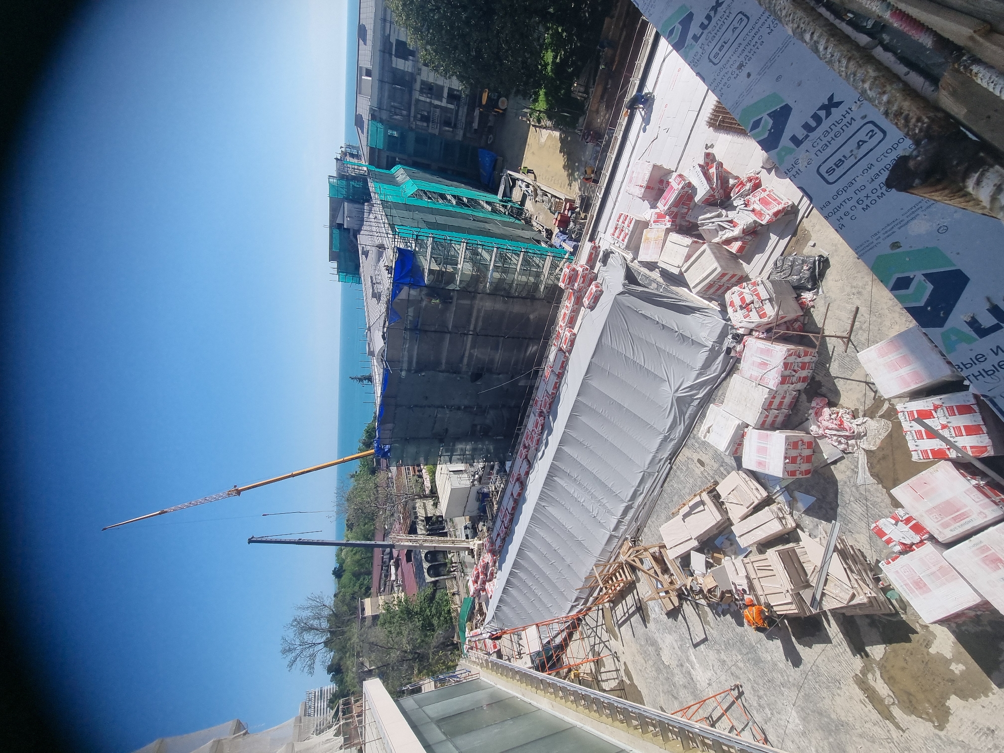 Here is a forensic description of the image:
**Location:** The image appears to be taken at a construction site in Sochi, Russia. This is inferred from the presence of construction materials labeled "LUX" and "SBL A2" in Russian, and the presence of what seems to be the Black Sea, typical of Sochi's geography.
**General Scene:** The photo shows a high-angle view of an active construction zone. In the background, what may be the Black Sea is visible, partially obscured by other buildings and construction infrastructure.
**Objects:**
* A large crane is visible in the distance.
* Buildings in various states of construction, with significant scaffolding.
* Stacks of materials appear to be construction components (possibly bricks or blocks).
* A large white tarpaulin or protective covering.
* Construction debris and wooden pallets.
* Construction material labeled with Russian text.
**People:**
* At least one person is visible at the bottom right, wearing what appears to be a safety helmet and/or construction gear.
**Actions and Interactions:**
* The visible individual is involved in work related to the construction materials, but the exact task is not clear.
* The crane indicates active lifting/moving of materials.
**Environment:**
* The scene seems to be taking place on a clear, sunny day.
* The surrounding area includes both existing structures and ongoing construction projects.
**Additional Observations:**
* The image may be taken from a higher elevation, potentially from another building under construction, or a higher floor of the building being constructed.
* The angle and framing of the photo suggest an informal, possibly opportunistic capture of the construction site.