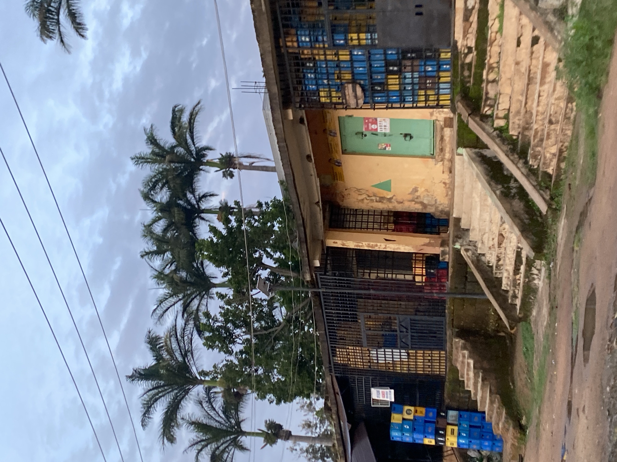 The image depicts a storefront in Jos, Nigeria, on a cloudy day. The building is made of concrete with a pale yellow facade, showing signs of wear and tear. A green metal door is visible in the center, with a small sign featuring red and white text above it. The storefront is protected by black metal bars. To the left of the door, a stack of blue and yellow plastic crates are piled high. To the right of the door, more blue and yellow crates are visible behind the bars.
The building is elevated from the street level, with two sets of concrete stairs leading up to it. The stairs are weathered and some have moss growing on them. The area in front of the building is a dirt road with puddles of water, indicating recent rain. Palm trees and other green foliage are visible behind and around the building, suggesting a tropical or sub-tropical climate. Power lines are strung across the sky. The overall impression is of a typical urban commercial setting in Nigeria.