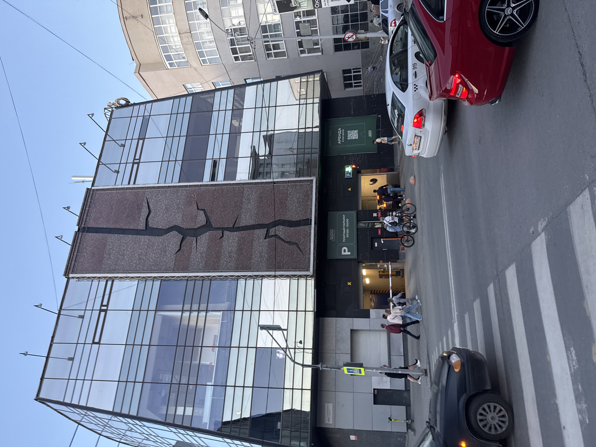 The image depicts a street scene in Yekaterinburg, Russia, in front of a modern, multi-story building with a glass facade and a large, textured vertical panel. In the foreground, a black car is stopped at a pedestrian crossing. Behind it, a white car and a red car are visible. Pedestrians are crossing the street. The building features a ground-level entrance to a parking garage, indicated by a barrier gate and signage. To the right of the parking entrance, a large green sign advertises "ARENDA" (rent) with a QR code. The sky is clear and blue. Several people are walking near the building entrance and on the sidewalk. Bicycles are also present. The overall scene is characteristic of a commercial or mixed-use area in a Russian city.