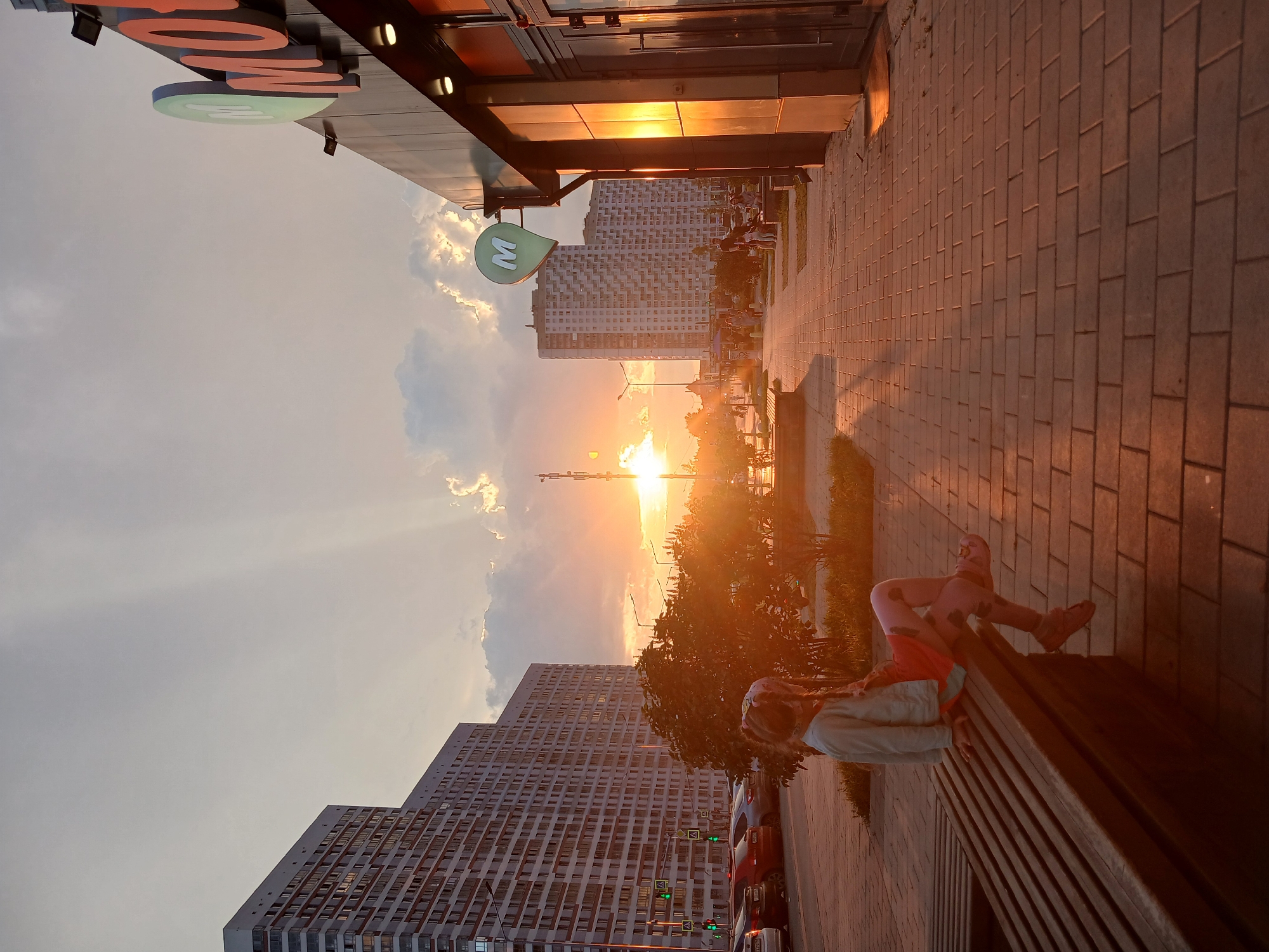 The image depicts a sunset scene on a street in Yekaterinburg, Russia. A young girl with braided hair sits on a wooden bench, facing away from the camera, looking towards the setting sun. She is wearing a light blue jacket, a pink shirt, and patterned pink leggings. The sun is low on the horizon, casting long shadows and creating a warm, orange glow that illuminates the scene.
In the background, tall, modern apartment buildings line the left side of the street. Cars are parked along the curb. A paved walkway extends into the distance on the right, leading to a building with a prominent "M" logo on its facade, indicating a commercial establishment, possibly a supermarket or a cafe. The sky is partly cloudy, with rays of sunlight breaking through, creating a dramatic effect. The overall atmosphere is tranquil and serene, capturing a moment of evening calm in an urban environment.