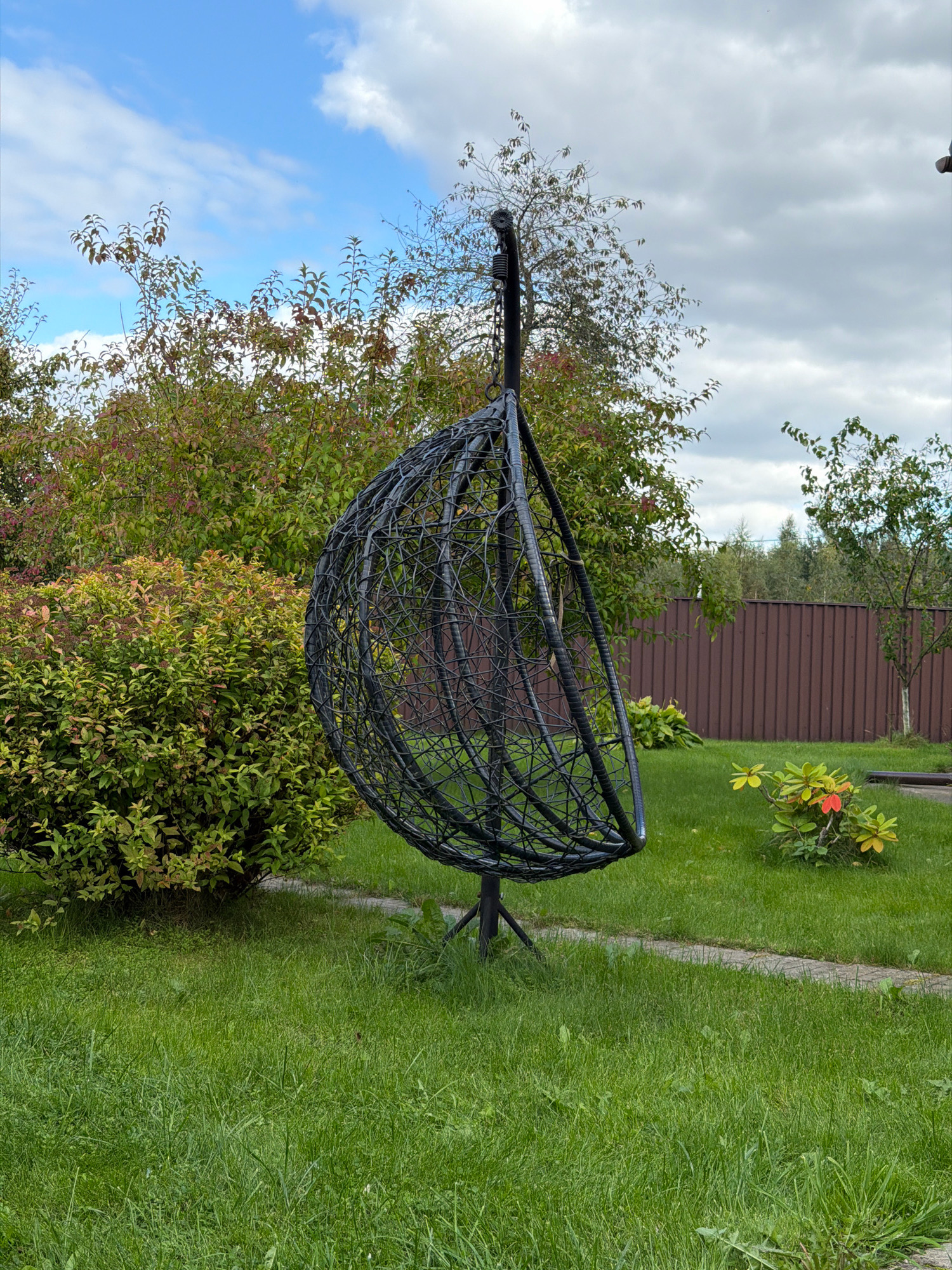 The image depicts a backyard scene in Бабініцкі сельскі Савет, Belarus. A black, woven hanging chair is centrally positioned on a stand in a grassy area. To the left of the chair is a large, dense green bush, and behind that, trees with green and reddish-brown foliage. Further back, a brown wooden fence runs horizontally across the image. Several trees are visible behind the fence. The ground is covered in green grass, with a light-colored paved path curving from the foreground towards the right. The sky above is a mix of blue with white clouds.