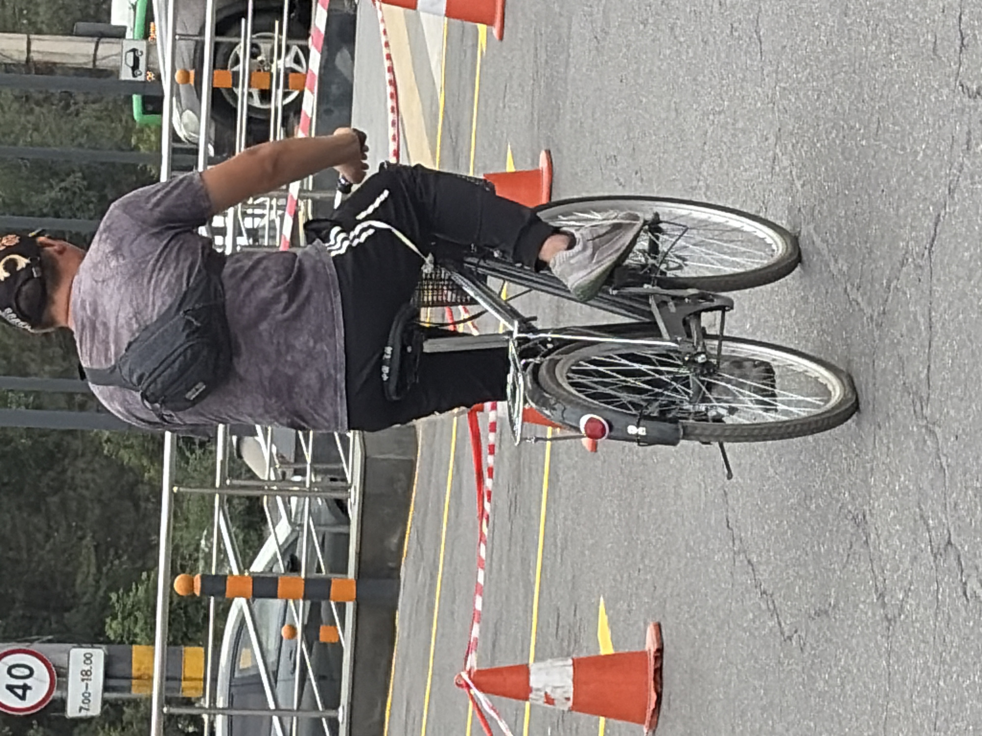A person rides a bicycle on an asphalt road. The person is wearing a gray t-shirt, black pants with white stripes, and a black fanny pack worn across the chest. The person is also wearing a black baseball cap and gray sneakers. The bicycle is black with a metal basket on the back. Orange traffic cones with red and white striped tape are visible in the background. A speed limit sign indicating 40 is posted on a pole. The scene appears to be in an outdoor urban environment, possibly a parking lot or street.