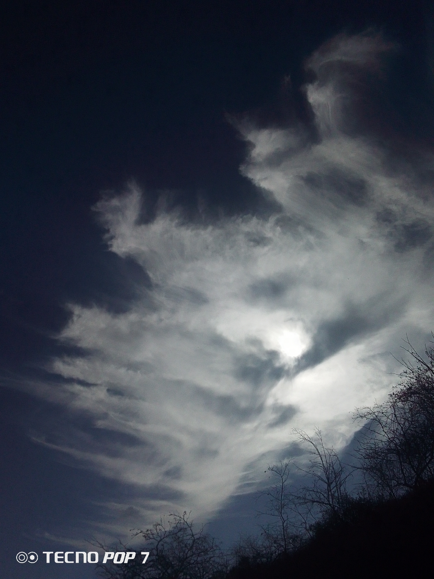 The image shows a sky with a mix of dark blue and clouds, taken in Altagracia, Venezuela. Some trees are visible against the sky on the right side and bottom of the picture, appearing dark against the light sky. The sky is brighter in the center, where the sun seems to be shining through the clouds. The overall image has a dark tone. The words "TECNO POP 7" are visible in the lower-left corner.