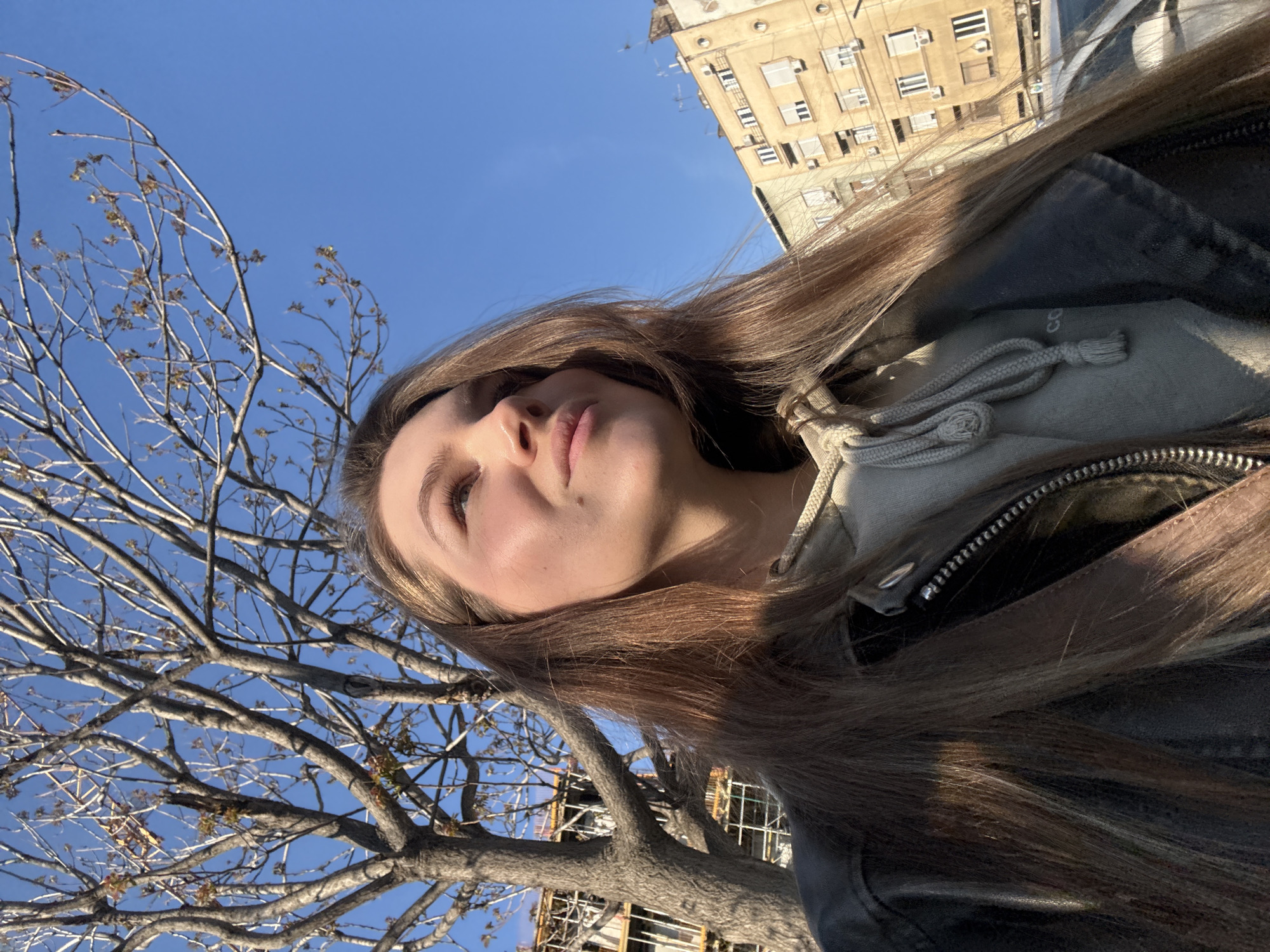 A young woman stands outside in Belgrade, Serbia. She is facing upward, with a leafless tree beside her. In the background, is a building with many windows. The sky is blue. The woman is wearing a grey hoodie under a dark jacket. Her hair is brown and long. Scaffolding can be seen among the tree branches.