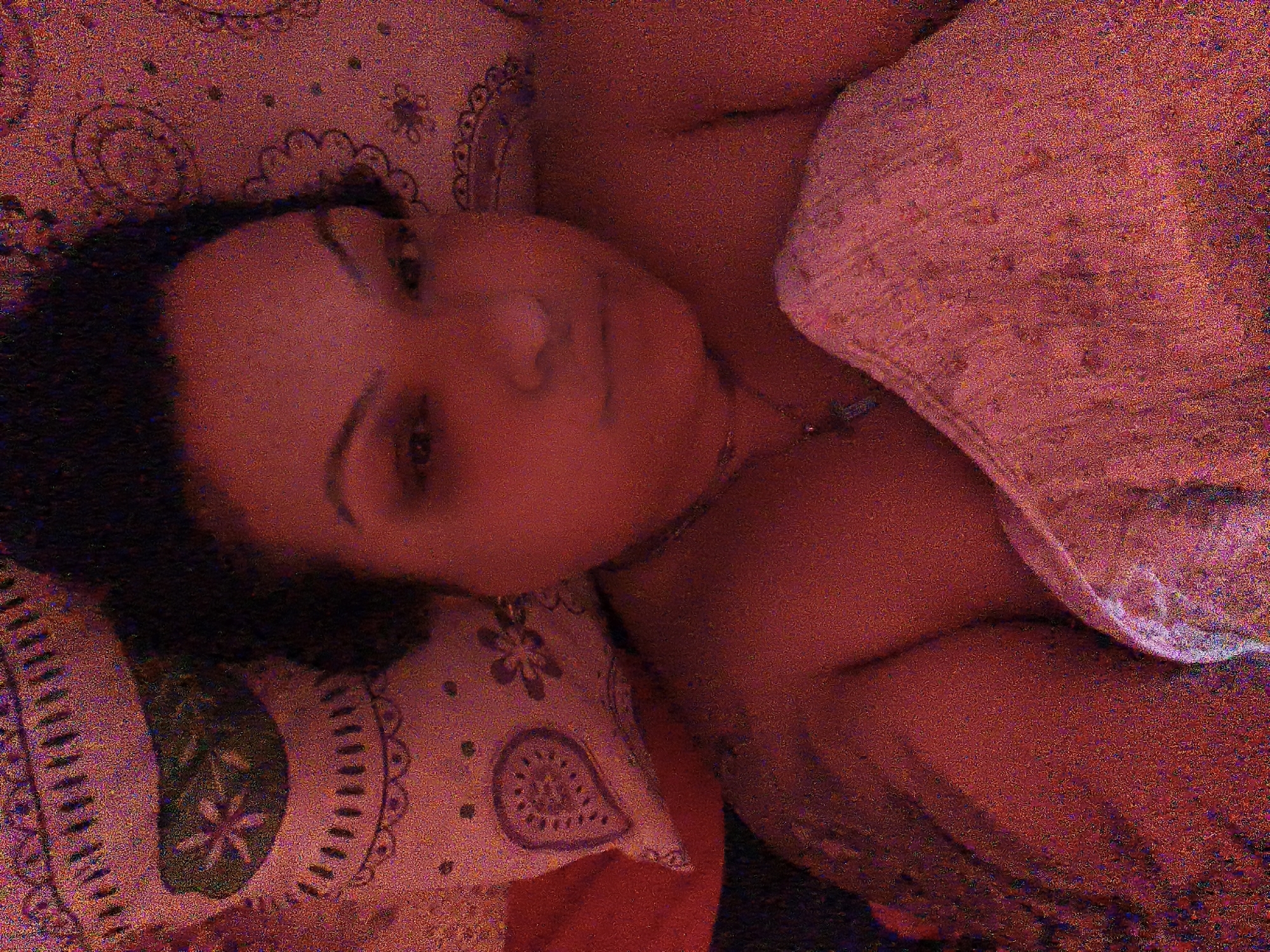 The image displays a close-up, sideways view of a person's face and upper body. The person has dark hair and appears to be lying down. They are wearing a light-colored, patterned garment draped over their shoulder and chest. The background is a reddish-pink textured surface with subtle, darker patterns resembling paisley or floral motifs. The lighting is dim and casts a warm glow over the scene. The image is grainy and has a low resolution, making fine details indistinct. There is no discernible information or context that places the image in the city of Komotini, Greece.
