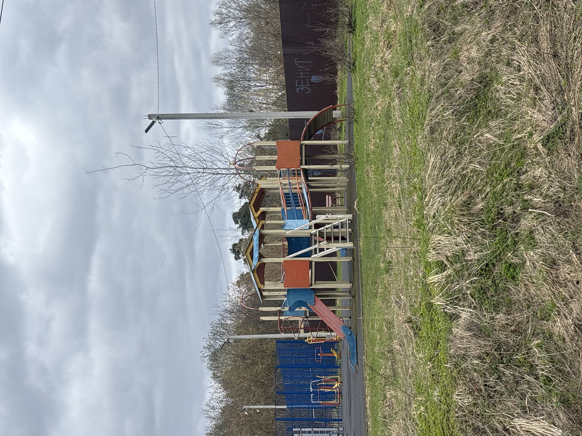 The image shows a playground in Razdolie, Russia. The playground equipment is multi-colored and features a slide, stairs, and platforms. The playground is bordered by a brown fence with Cyrillic graffiti and a blue metal fence. Overgrown grass surrounds the playground. Utility poles and bare trees are visible. The sky is overcast.