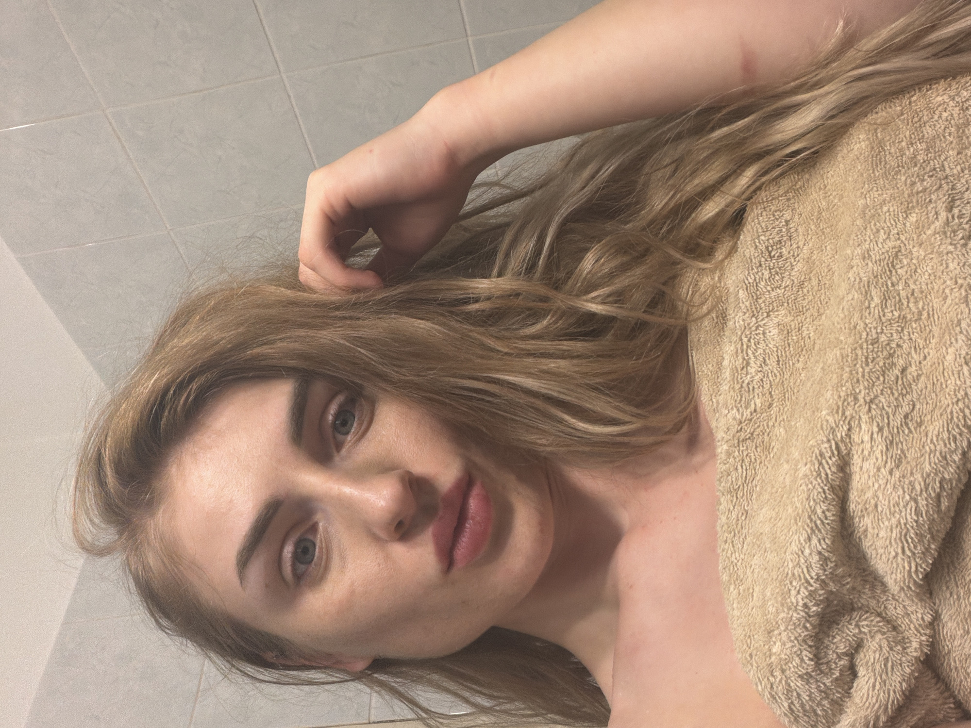 The image shows a close-up, selfie-style shot of a young woman with blonde hair and blue eyes. She is wrapped in a tan towel around her chest and is looking towards the camera. Her left hand is raised and her fingers are in her hair, near her temple. The background appears to be a tiled wall, suggesting a bathroom environment. The lighting is somewhat dim and the image has a slightly grainy quality. There are no discernible landmarks or features that would indicate a specific location within a city.