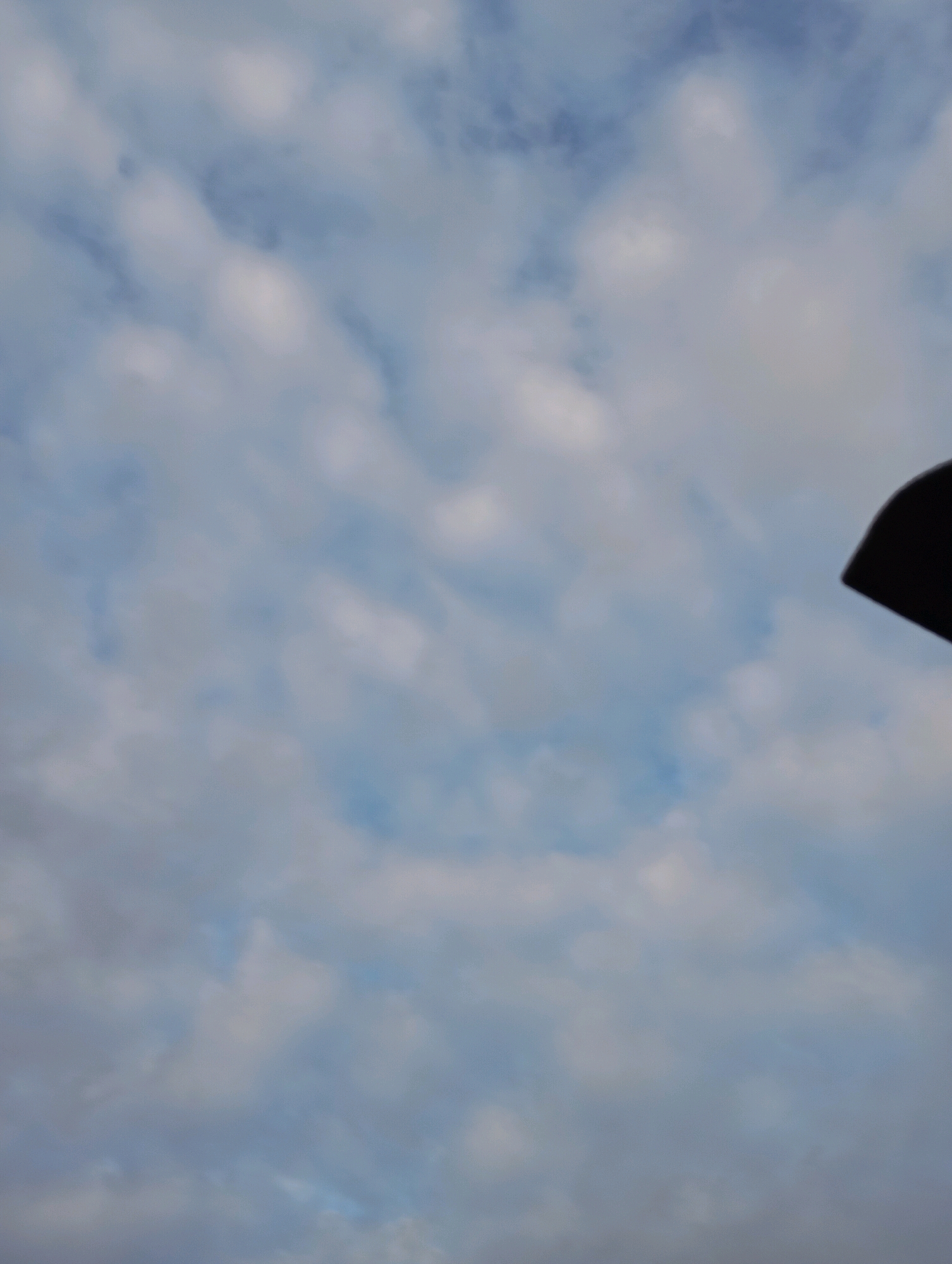 The image captures a cloudy sky above Horishni Plavni, Ukraine. A portion of a dark, curved object, possibly a part of a structure or equipment, is visible in the upper right corner of the frame. The rest of the image is dominated by a soft blue sky filled with scattered white clouds.