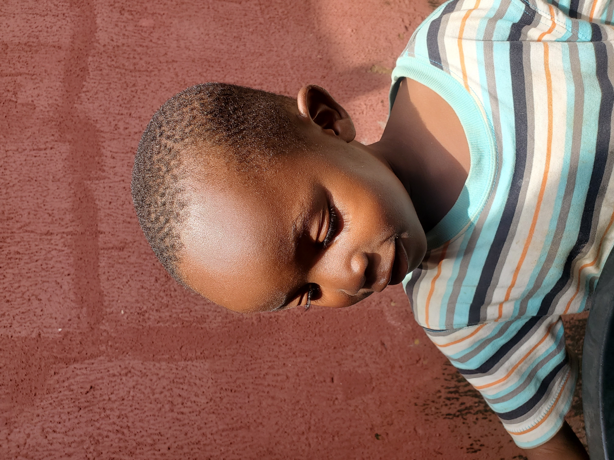 The image features a close-up, horizontally oriented shot of a young child from the chest up, resting on a reddish-brown, textured surface. The child has dark skin and short, textured hair. Their eyes are closed, and their head is tilted to the side, suggesting they are sleeping or resting. Sunlight casts a shadow across the child's forehead and cheek, indicating it is daytime, likely outdoors due to the natural lighting and the textured surface which could be a ground or wall.
The child is wearing a t-shirt with a colorful striped pattern, featuring alternating bands of light blue, white, gray, navy blue, and orange. The collar of the t-shirt is light blue. The background is entirely composed of the reddish-brown textured surface, with no other distinct objects or elements visible besides the child and their clothing. No text is visible in the image. The overall mood conveyed is one of peaceful rest. The location context provided is Jos, Nigeria.
