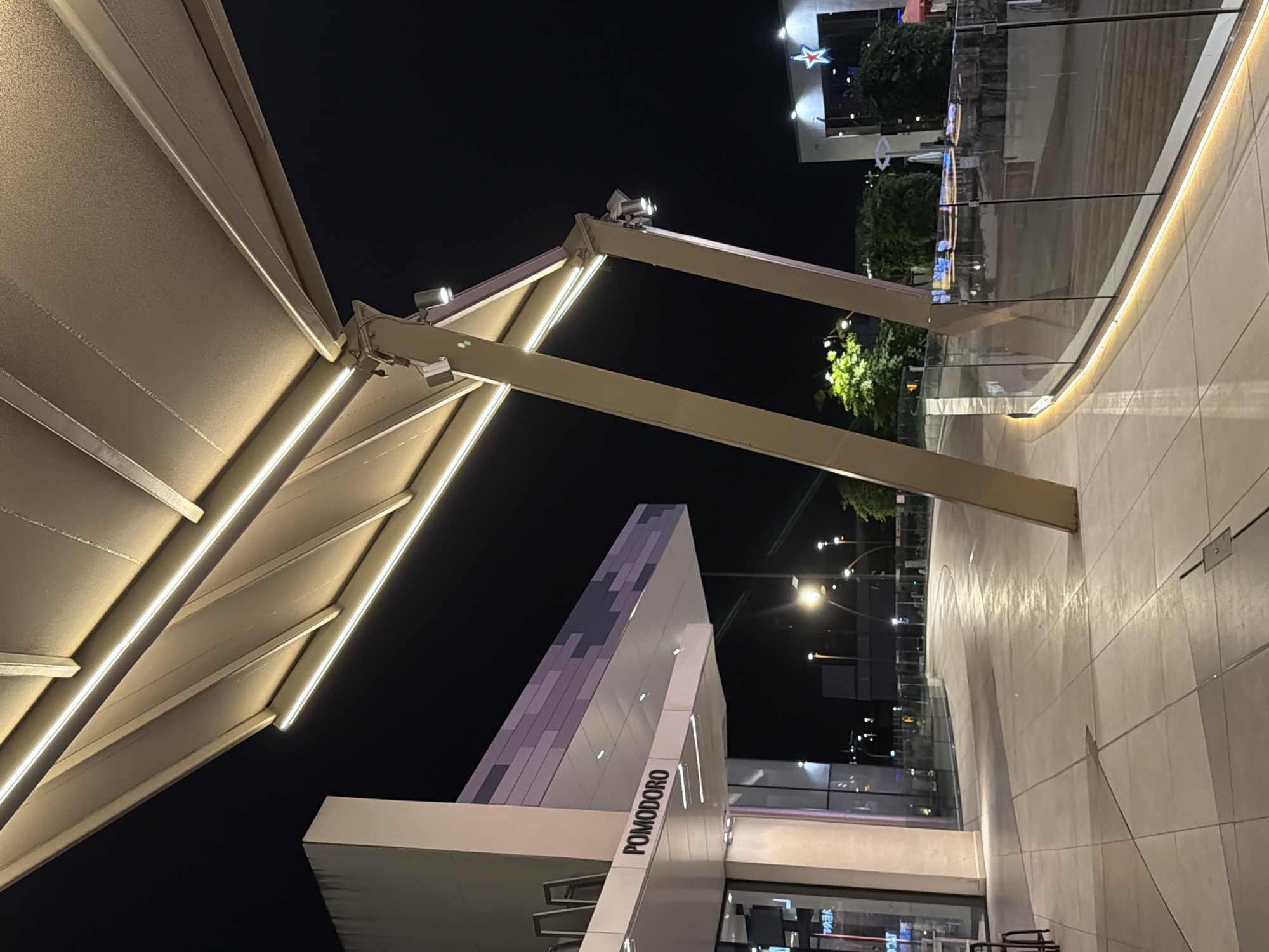 This media file captures an outdoor scene at night, illuminated by artificial lighting. The foreground is dominated by a large, angled awning supported by metallic beams. The awning itself is lit from beneath by bright, linear LED lights. In the mid-ground, a building with a modern, angular design is visible, featuring a textured, multi-toned facade. A sign on this building clearly reads "POMODORO".
Beyond this, the scene opens up to a walkway paved with large, light-colored tiles. A glass barrier separates the walkway from a lower level or area, also illuminated. Several streetlights are visible in the background, casting pools of light. Trees with lush green foliage are scattered throughout the background, appearing vibrant despite the darkness. In the upper background, building structures and more lights suggest an urban or commercial setting, possibly a shopping area or entertainment district. The overall mood is calm and inviting, with the lighting creating a sense of ambiance. The time of day is clearly night. There is no indication of weather conditions. There are no people visible in the media file.