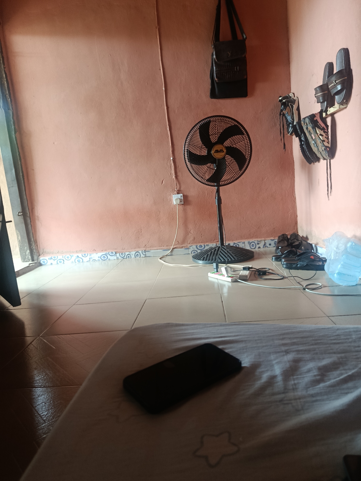 The image depicts an indoor scene, likely a bedroom or living area, in Ukpor, Nigeria. The walls are painted a peachy-pink color, with a blue and white tiled border running along the lower portion. The floor is tiled with light-colored squares.
Prominently featured is a black oscillating fan with a circular base and a black oscillating fan head with a yellow logo. The fan is positioned on the floor, connected to a power strip that is also on the floor. The power strip has multiple outlets and is plugged into a wall socket. Several electronic devices, possibly chargers or adapters, are plugged into the power strip, with various cables extending from them.
On the wall above the fan, a dark leather messenger bag with a flap closure is hanging. To the right of the fan, a shelf is mounted on the wall, holding two pairs of sandals and what appears to be a pair of athletic shoes. Further to the right, a large clear plastic bottle of water is placed on the floor.
In the foreground, a dark-colored smartphone lies on a white or light-colored fabric, possibly a bedsheet, which has a faint star pattern. The fabric is rumpled, suggesting it is a bed or sofa. To the left of the frame, a portion of a dark object, possibly a door frame or furniture, is visible. The lighting suggests it is daytime, with light streaming in from an unseen window, casting shadows on the floor and the fabric in the foreground.