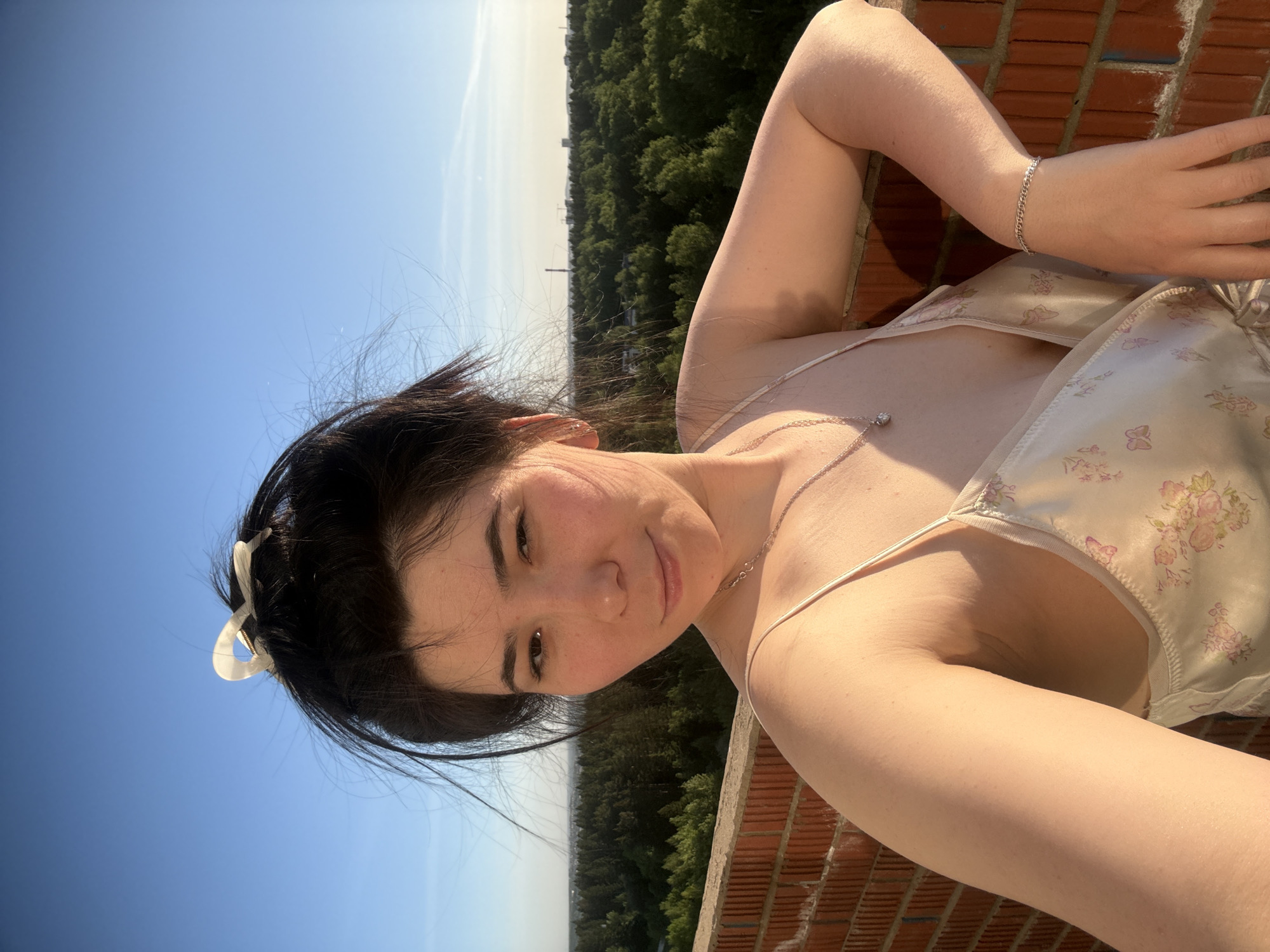 A young woman with dark hair pulled back by a white hair clip is taking a selfie outdoors. She is leaning against a brick structure. She is wearing a light-colored floral print camisole, a delicate necklace, and a silver bracelet. In the background, there is a dense green forest skyline, with a clear blue sky visible above. Based on the skyline, this is in Izhevsk, Russia.