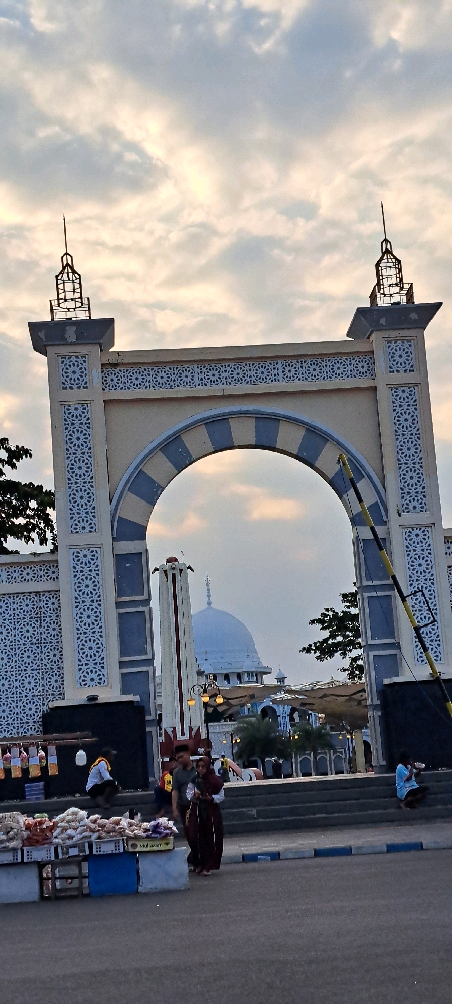 The image shows an arched gateway with Islamic architectural motifs, likely leading to a mosque or religious complex in Pasuruan, Indonesia. The sky is filled with cloudy, diffused light, suggesting either early morning or late afternoon.
In the foreground, a market stall is set up on the left, displaying various packaged goods and snacks. Several people are present. On the left, a person wearing a yellow shirt is crouching near the stall. In the center, three individuals are walking towards the right, with one person in the foreground wearing a dark maroon hijab and dress. To the far right, a person in a light blue shirt is sitting on steps.
Beyond the archway, a white mosque dome is visible, along with other structures that appear to be part of the religious complex. A tall, slender monument with red and white detailing stands to the left of the mosque dome. Trees are visible scattered throughout the background. The overall scene captures a blend of daily life with religious architecture, suggesting a public space in Pasuruan.
