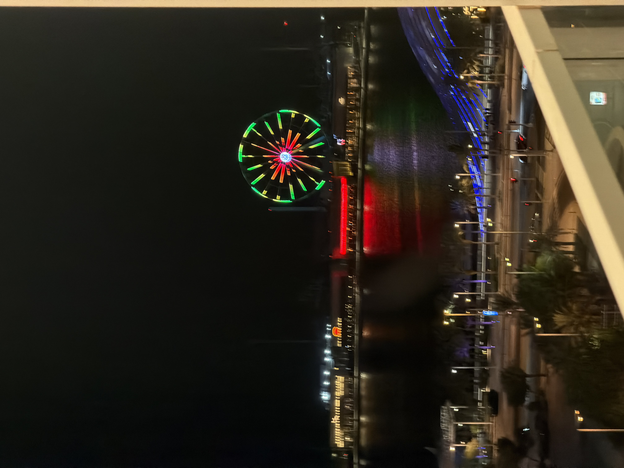 The image presents a nighttime, elevated perspective of a vibrant urban waterfront. In the immediate foreground, a white balcony railing with a glass panel is visible on the lower right, framing the scene. A dark body of water occupies the midground, reflecting the numerous bright lights from the opposite shore.
Dominating the midground-left is a large Ferris wheel, brightly illuminated with a distinct pattern: segments of green lights on its outer rim and radial spokes featuring red, yellow, and some blue lights converging towards a prominent blue central hub. Across the water, a series of illuminated buildings line the shore, appearing to be commercial establishments or restaurants, with their lights reflecting as streaks of color on the water's surface. A long structure to the left of the Ferris wheel emits a strong red glow, also reflected in the water.
Further back, a multi-lane road runs parallel to the waterfront. It is illuminated by numerous streetlights emitting a warm yellow-orange light. Several vehicles, including a dark car, are discernible on the road. Palm trees are scattered along the roadside and surrounding areas. On the far right, a distinctive architectural structure is illuminated with parallel blue lines, possibly a pedestrian bridge or a decorative element, with a few blurry figures, presumably people, visible on it. The sky above is completely dark, underscoring the artificial illumination of the scene.
Based on the distinctive Ferris wheel design (Eye of Yas), the architectural style, and the waterfront promenade, the location is identified as Yas Bay Waterfront on Yas Island, Abu Dhabi, UAE.