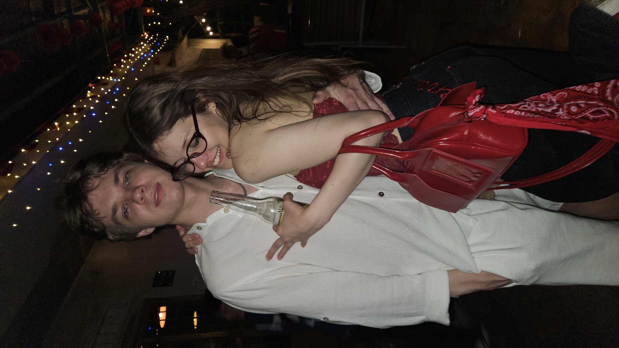 A woman with long brown hair and glasses embraces a man with blond hair. The woman wears a red top and black bottoms, and a red purse with a red bandana is on her shoulder. The man wears a white shirt and holds an empty Corona beer bottle. The setting appears to be a dimly lit outdoor area with string lights. The location is not discernible as Nha Trang, Vietnam from visual cues within the image.
