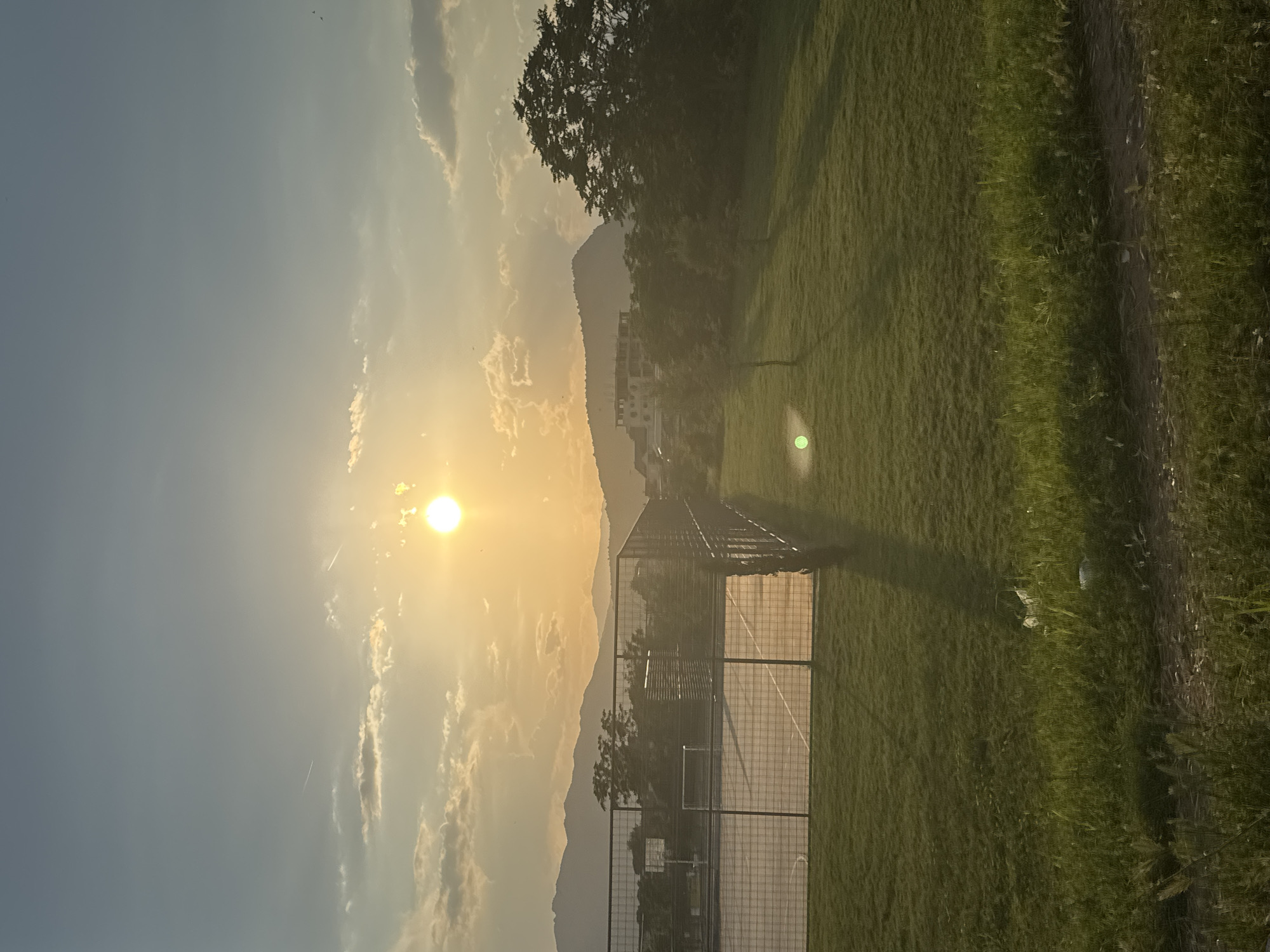 The image shows a landscape with a bright sun in the sky illuminating the scene. A metal fence encloses what appears to be a court or field with green grass. In the background, there are hills or mountains and a building. The location seems to be an open, possibly suburban or rural, area. The exact city is unknown.