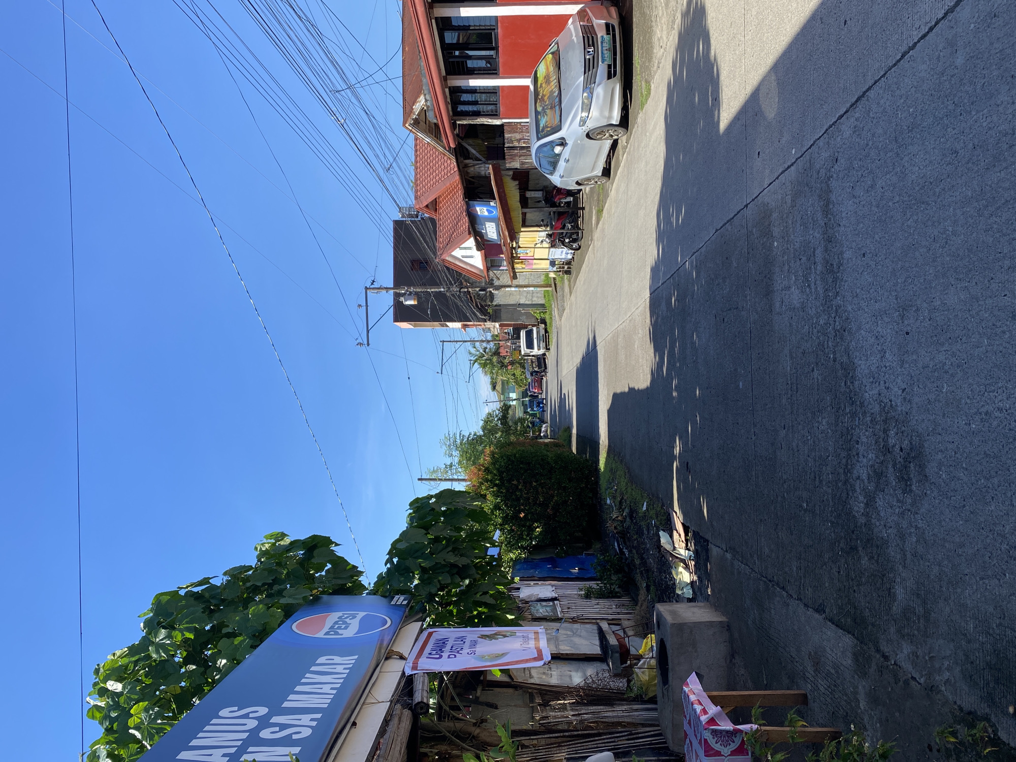The image shows a street scene in Davao City, Philippines on a clear, sunny day. The street is paved and appears to be a local road, with houses and small businesses lining both sides. A silver sedan with a Philippine license plate is parked on the right side of the street. In the foreground on the left, there is a table with a red and white checkered cloth and some plants. Above the table is a blue sign with white text and a Pepsi logo. Further down the street on the left, there are various structures, including a building with a red roof and some vegetation. On the right side of the street, there are several low-rise buildings with red roofs, typical of residential and small commercial establishments. The sky is a clear blue, with a network of power lines crisscrossing overhead. The overall impression is a typical urban street in the Philippines, with a mix of residential and commercial activity.