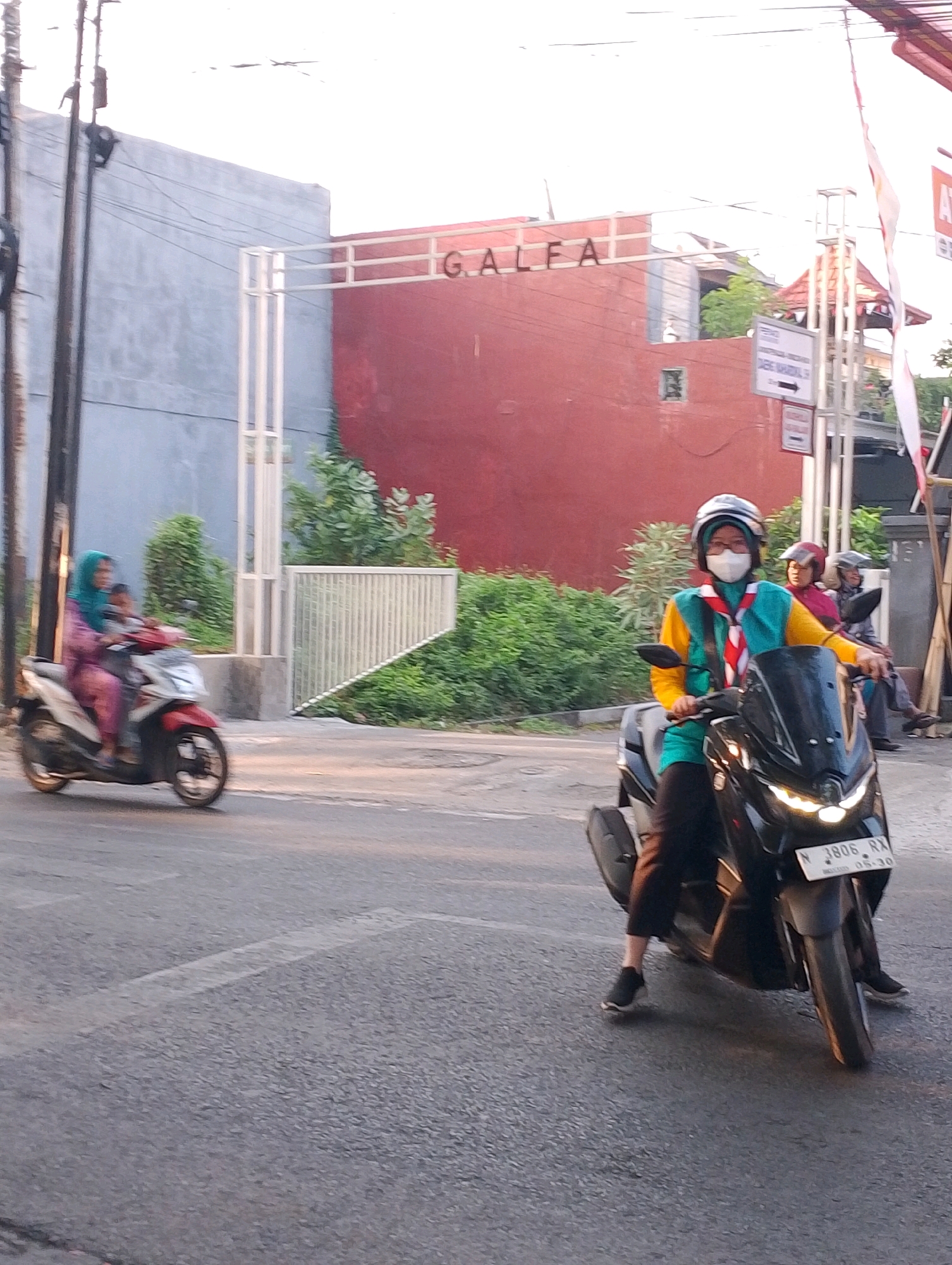 The image captures a street scene in Probolinggo, Indonesia, with a focus on two motorcycles on a paved road. In the foreground, a person wearing a scout uniform, helmet, and face mask rides a black scooter. The scout uniform consists of a yellow long-sleeved shirt with a teal vest and a red and white scarf. The scooter has a license plate that reads "N 3806 RX". Behind this scooter, another scooter is partially visible with two individuals on it. The person in front wears a turquoise hijab and pink attire, while a child is seated behind them.
The background features a building with red walls and a sign that appears to say "GALFA". There are also white metal fences and lush green foliage. The sky is bright, suggesting it is daytime, possibly late afternoon or early morning due to the warm lighting. The overall impression is that of a typical urban street in Indonesia.