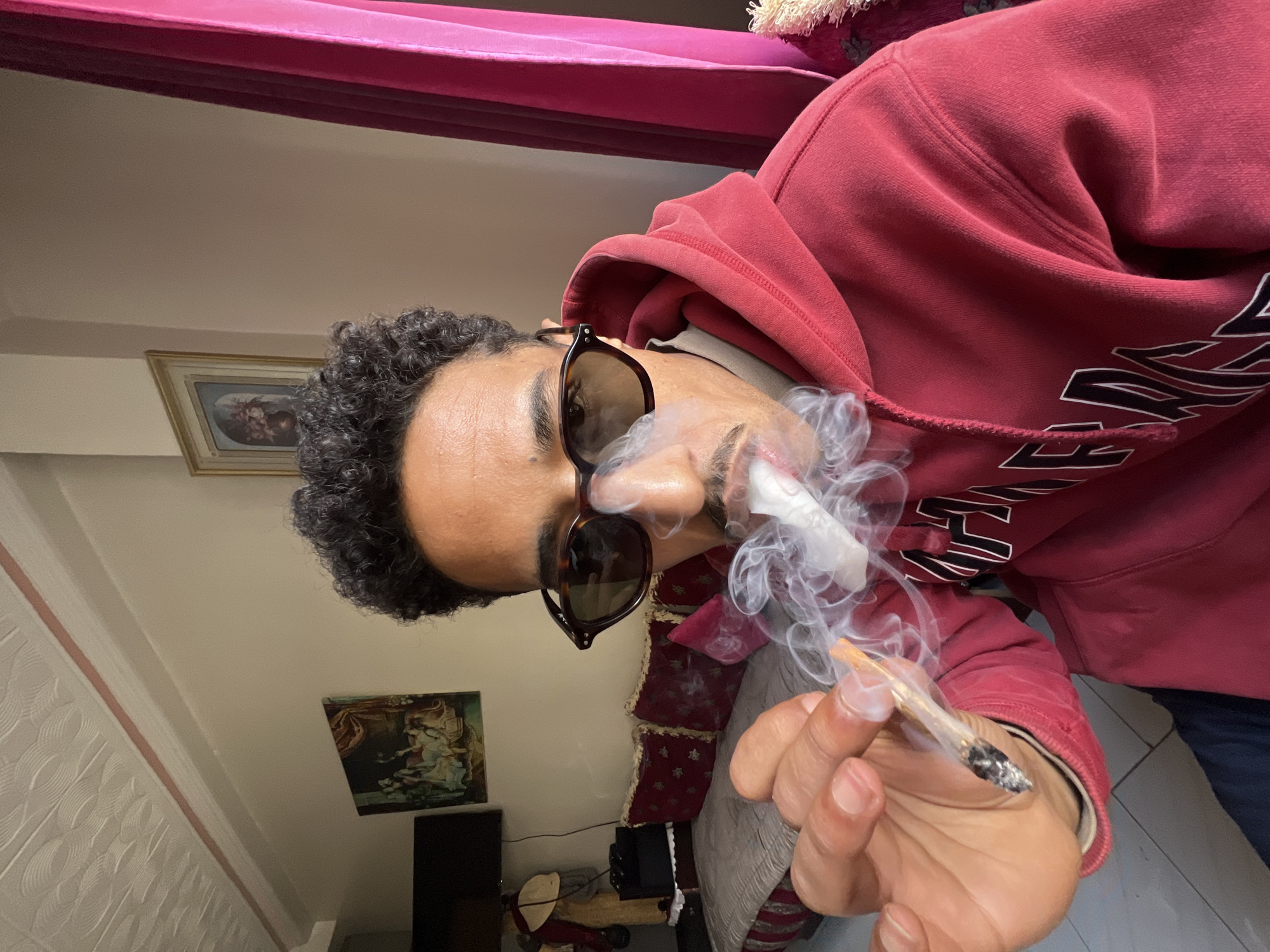 A man with dark curly hair and sunglasses is holding a lit marijuana cigarette (joint) and exhaling smoke. He is wearing a red hooded sweatshirt. The background includes framed artwork on a wall, a red curtain or upholstered furniture trim, and white textured wall paneling. It is impossible to determine the location in Tanger, Morocco, based on this image.