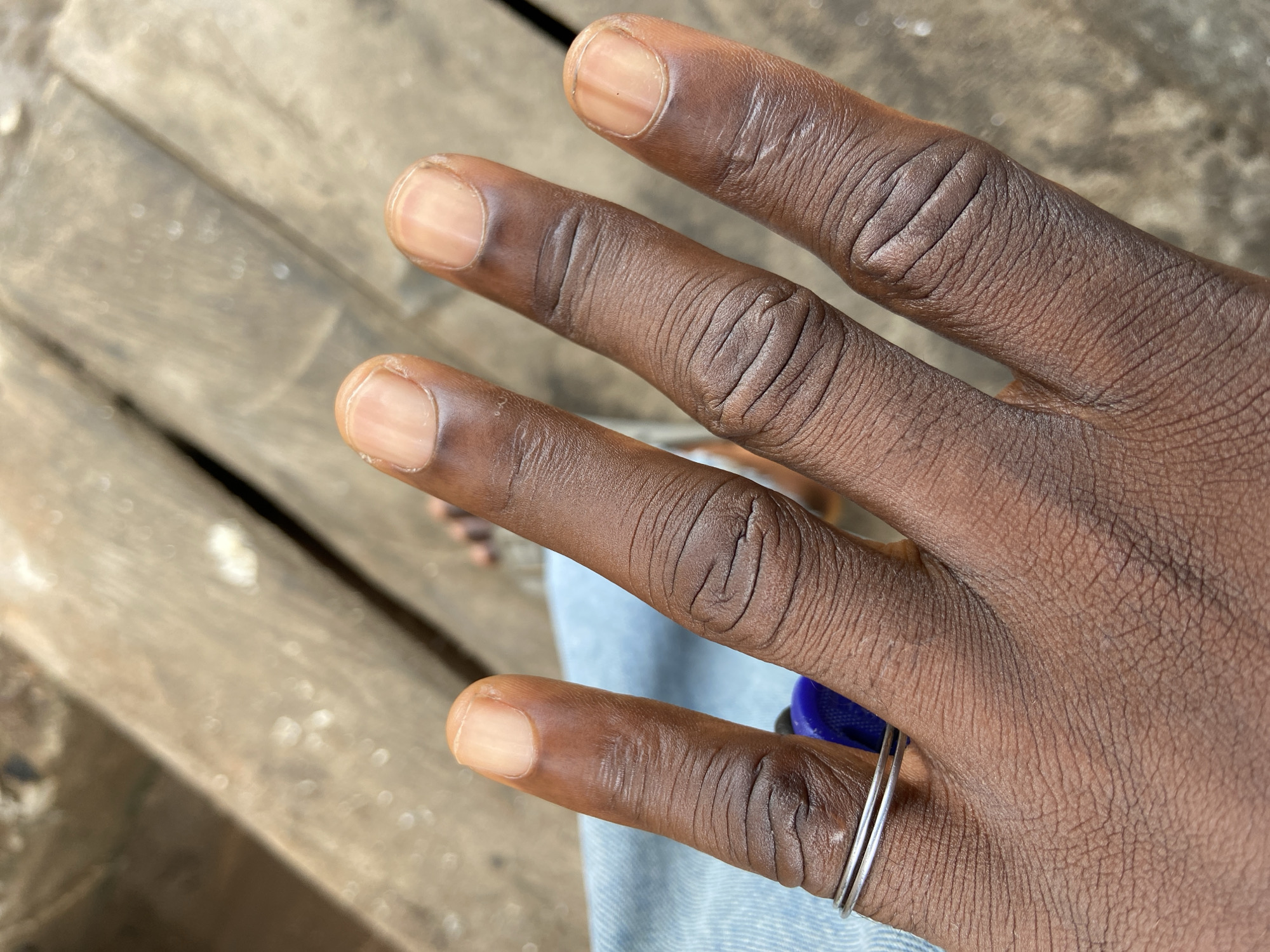 A close-up shot depicts a brown-skinned human hand with short, unpolished nails. The hand has visible wrinkles and fine lines. On the ring finger, there are two thin, silver rings. The hand rests on a pair of light-blue denim jeans. The background includes a blurred wooden surface with varying shades of gray and brown. No specific landmarks or objects pinpoint a location in Mararaba, Nigeria.
