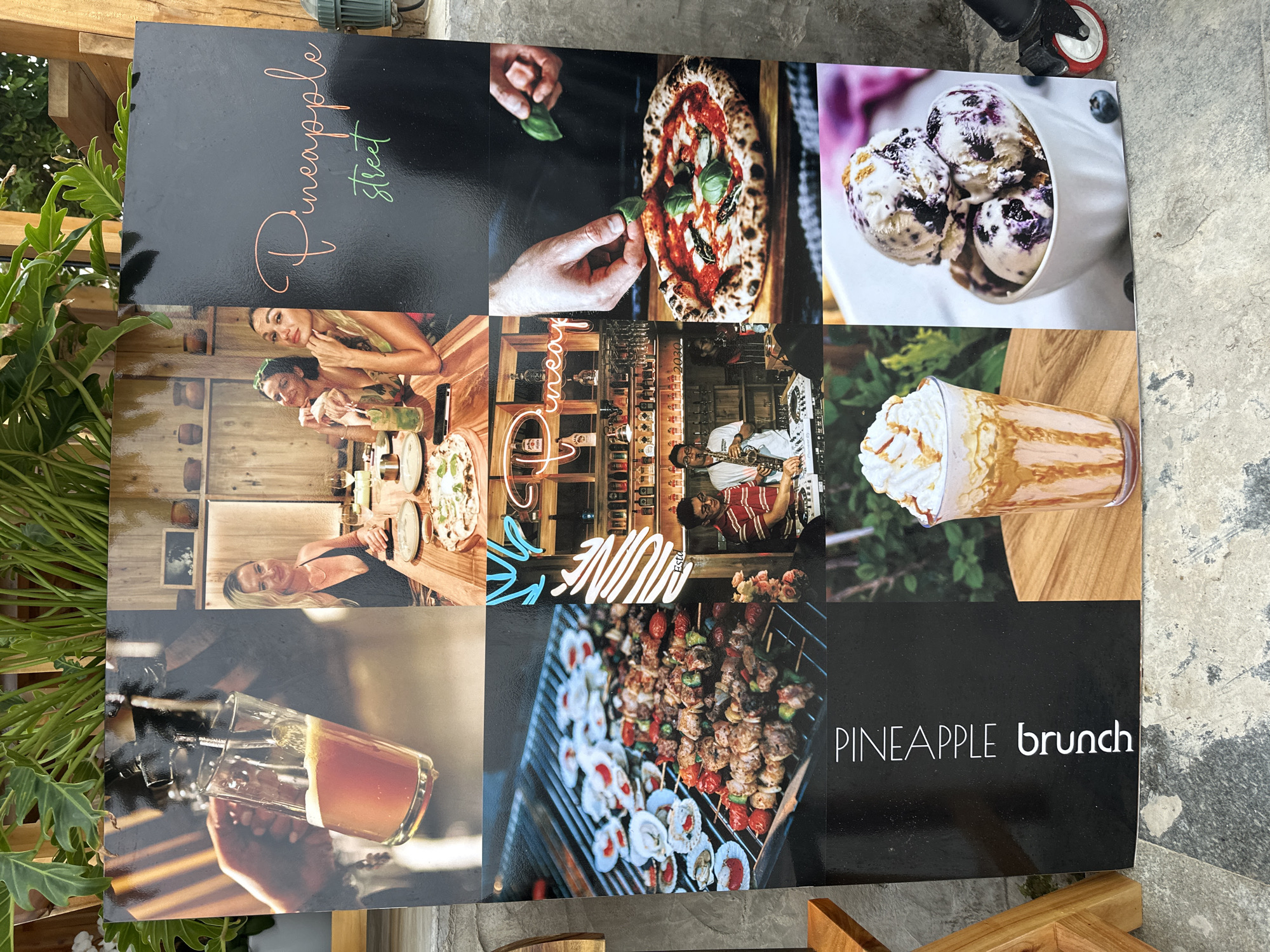 The image is a photograph of a large sign advertising "Pineapple Street" and "Pineapple Brunch" in Phu Thuy Ward, Vietnam. The sign is composed of a grid of nine smaller images showcasing various aspects of the establishment.
The top row features:
* An image of a hand pouring beer into a glass.
* Two women dining at a table, with pizzas and drinks visible.
* The "Pineapple Street" logo in script on a black background.
The middle row displays:
* Grilled food, including skewers and scallops, on a barbecue.
* A neon sign reading "Pineapple" over shelves stocked with bottles.
* A close-up of a pizza being garnished with basil leaves.
The bottom row shows:
* A DJ playing music while a man plays the saxophone.
* The text "PINEAPPLE brunch" vertically on a black background.
* A tall glass filled with a creamy drink topped with whipped cream and caramel drizzle.
* A bowl of blueberry ice cream with blueberries scattered around it.
The sign is placed outdoors, leaning against a grey wall with some greenery above it and a wooden chair leg visible on the left. The ground appears to be tiled concrete.