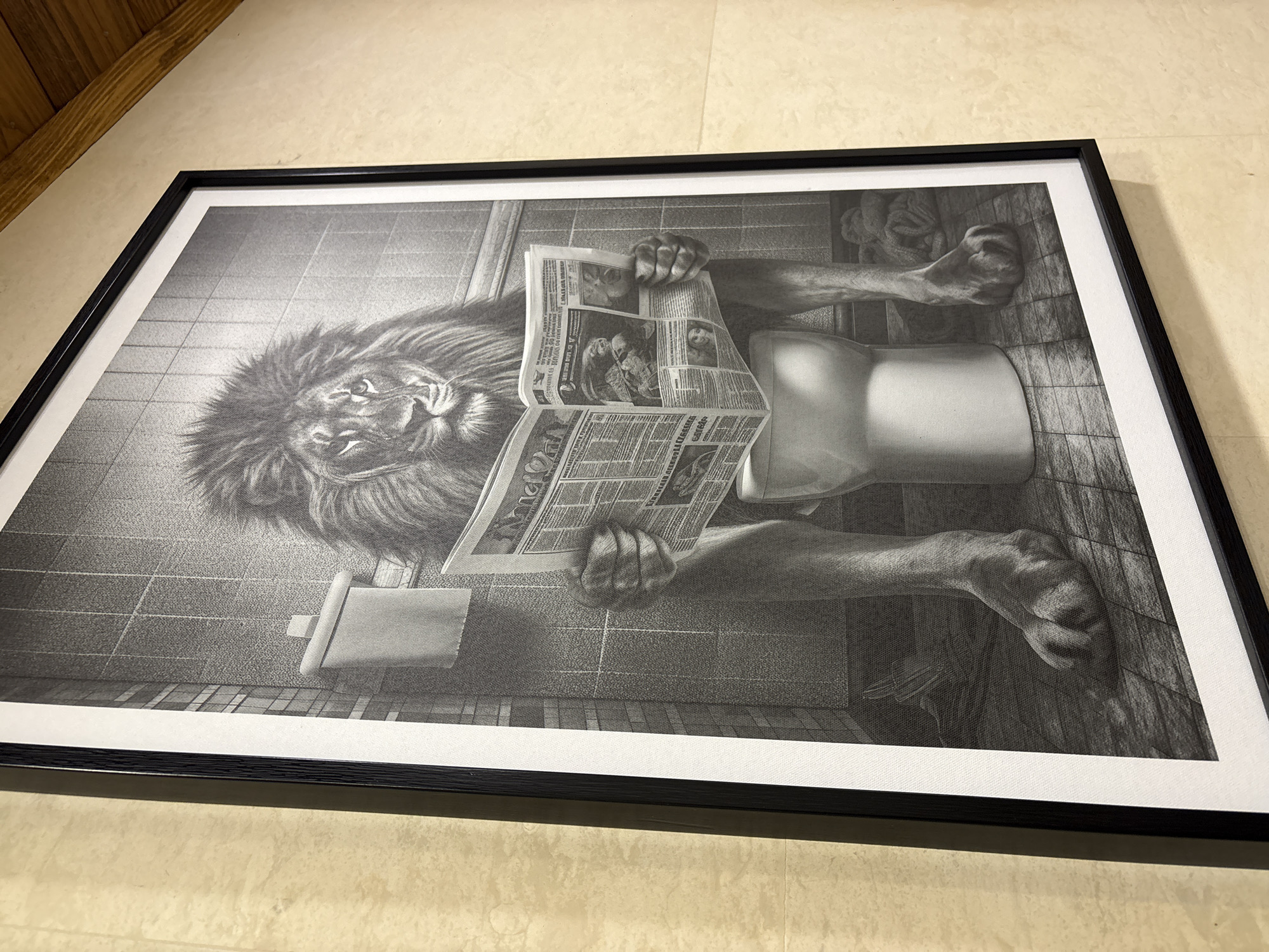 A framed black and white print depicts a lion seated on a toilet reading a newspaper. The scene is a bathroom setting, evident from the tiled walls, toilet paper dispenser, and toilet. The artwork is framed with a black frame and a white border, and it is placed on a surface that appears to be a tiled floor. There are no indications that the image was taken in Koprivlen, Bulgaria.