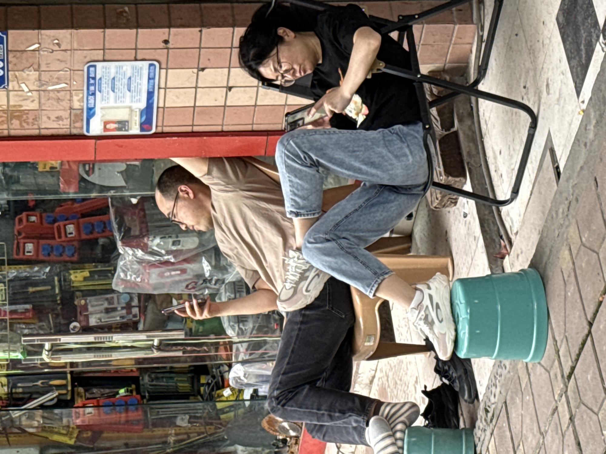 A street scene in Chongqing High-tech Zone, China, shows two people in front of a shop window with a display of tools and equipment. A man in a tan shirt and black pants stands with his right arm extended, holding a phone. He wears striped socks and off-white sneakers. A woman sits on a folding chair, wearing a black t-shirt, blue jeans, and white sneakers. She appears to be handling money. They are positioned on a sidewalk with patterned pavement. A blue sign is visible on the wall above the shop window. One of the man’s feet rests on an upturned green bucket. A pair of black shoes lie on the sidewalk beneath the woman's feet.