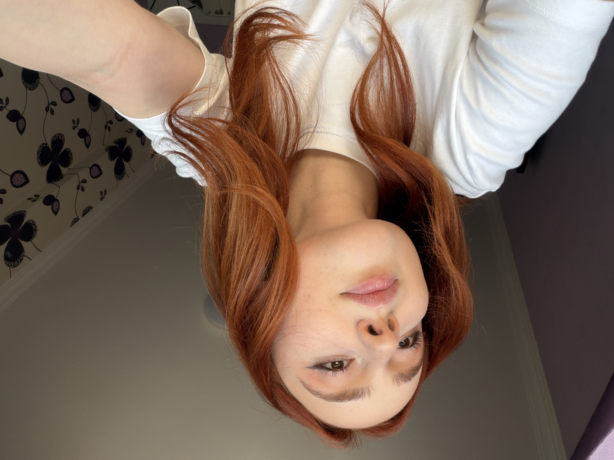 The image shows a woman with long, auburn hair wearing a white t-shirt. She is positioned with her head tilted downwards, creating an unusual perspective for the viewer. Her face is prominent, displaying her features such as her eyes, nose, and lips clearly. The background includes a wall with a floral wallpaper pattern in the upper left, a plain grey wall, and a dark, undefined surface. No information in the image suggests the location is Tbilisi, Georgia.