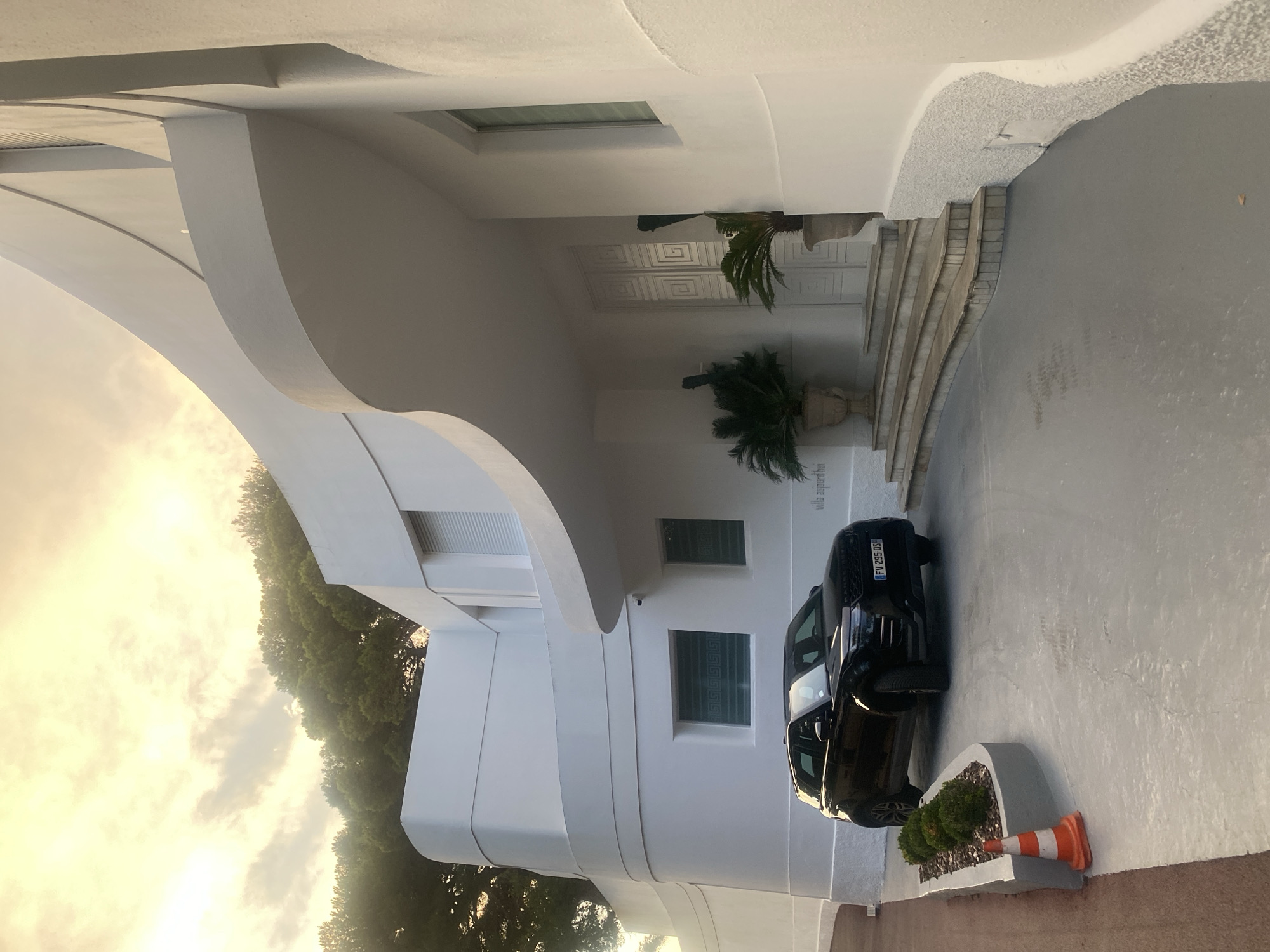 The image shows the exterior of a modern white villa with curvilinear architecture, located at Villa Augure, 7 Avenue Saint-Lazare, 06160 Antibes, France. A black Range Rover Evoque is parked on a light gray paved driveway in front of the villa. The car has a French license plate that reads "FV-295-DS". Two potted palm plants flank a set of stone stairs leading to the villa's entrance. The sky above is filled with soft, warm-toned clouds, suggesting either sunrise or sunset. The environment is lush with green trees visible in the background. A traffic cone is placed on the edge of the driveway. The overall scene is one of luxury and tranquility.
