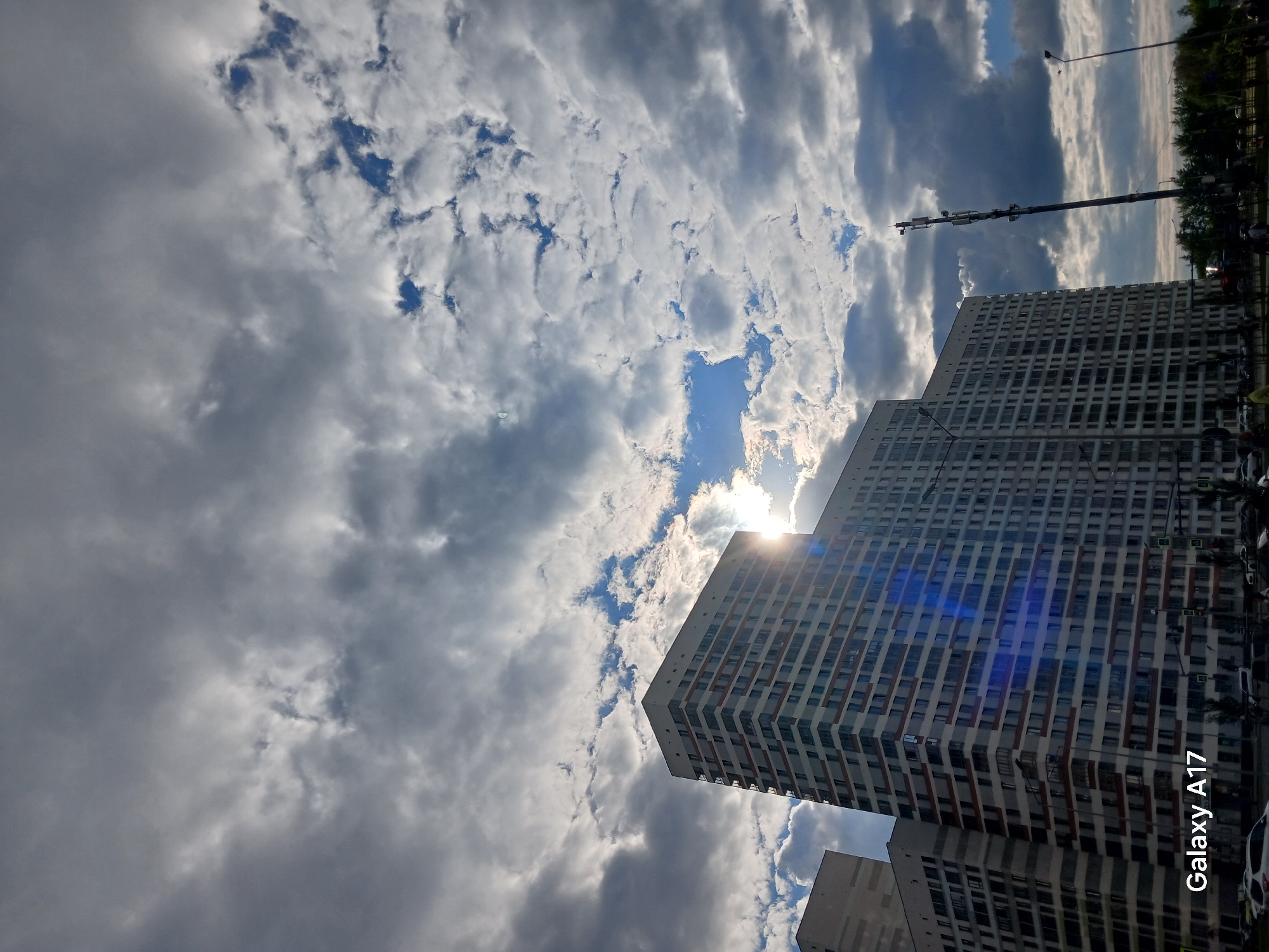 This is a low-angle outdoor shot depicting a modern urban landscape under a partially cloudy sky. The image captures several tall, contemporary high-rise apartment buildings from below, emphasizing their height against the expansive sky. The buildings are predominantly light-colored with numerous rectangular windows, some featuring darker horizontal accents.
The sky is a dominant feature, filled with a dynamic mix of white and grey clouds. Patches of clear blue sky are visible, especially towards the upper middle and near the sun. The sun is partially obscured by clouds but breaks through, creating a bright lens flare or starburst effect over the upper right facade of the tallest building, indicating a bright daytime setting, likely late morning or afternoon.
To the left of the main buildings, a slender communication tower or utility pole with multiple antennae stands tall against the sky. At the very bottom right of the image, a strip of green trees and hints of a road with parked cars are visible, providing a glimpse of ground-level activity in this urban environment, consistent with a setting in Yekaterinburg, Russia.
No specific human activity is depicted. The overall impression is one of a bright, yet atmospheric day with varying cloud cover. The text "Galaxy A17" is visible in white, oriented vertically along the bottom right edge of the image, identifying the camera used.