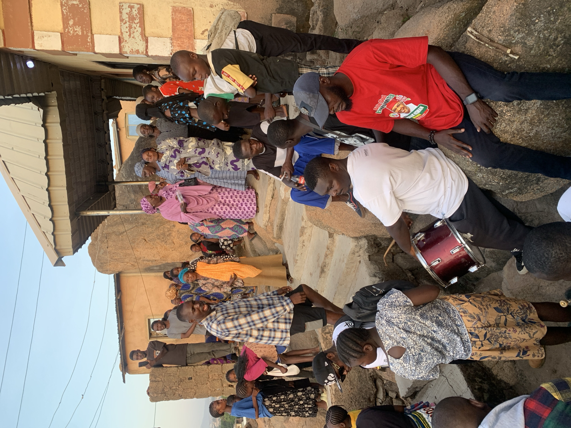 This media file captures a lively outdoor gathering of a diverse group of people, including men, women, and children, in what appears to be a village or communal setting. The overall scene suggests a social event or celebration.
**Subjects:** The image features numerous individuals, many looking towards a central activity, though the exact focus is not entirely clear. Men, women, and children are present, dressed in a mix of casual attire (t-shirts, jeans) and vibrant traditional patterned clothing. One man in a red t-shirt is prominently seated on a large rock, while another man holds a red drum, suggesting a musical or ceremonial aspect to the gathering. Several other men are standing or seated around.
**Objects:** A notable object is the red drum with silver hardware, held by a person on the right side of the image. A man in a blue shirt holds a Coca-Cola bottle. Large, light-colored rocks form part of the terrain, acting as natural seating for some attendees. In the background, simple buildings with corrugated metal roofs are visible, indicating a residential area.
**Activity or event taking place:** The crowd is gathered closely, with some individuals standing and others seated on the rocks or ground. The presence of the drum strongly suggests music, performance, or a traditional ceremony is either underway or about to begin. People appear engaged and observant, with some smiling faces visible. It seems to be a community gathering or a public event.
**Notable details:** The lighting indicates late afternoon or early evening, with a clear, bright sky. The setting appears rustic, characterized by unpaved ground, natural rock formations, and simple architecture. Emotions conveyed are generally positive and engaged. A man in the red t-shirt is wearing a smartwatch.
**Visible text:** Text is visible on a few items:
* On the red t-shirt, the words "NGAJI EXPRESSION" are prominent, along with an image of a man and "TOBI OGUNSAN", suggesting political or promotional merchandise.
* A white t-shirt bears the brand "LANCIOT".
* A dark t-shirt with yellow sleeves has "LOVE LIFE" printed on it.
* The Coca-Cola bottle has its brand name visible.