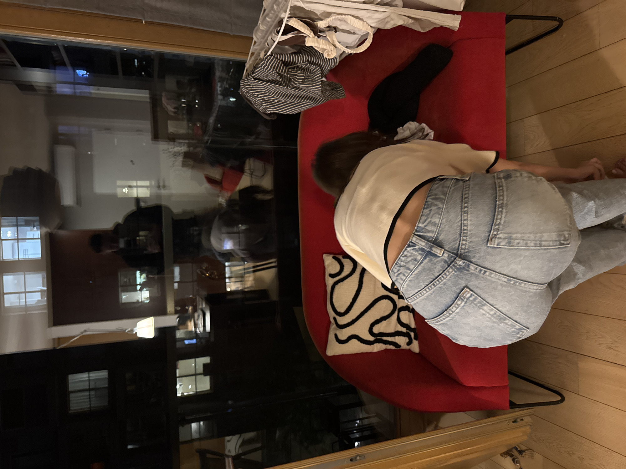 The photograph shows a person lying prone on a red chaise lounge. The person is wearing light blue denim jeans and a cream-colored top with black trim. A patterned pillow rests behind the person. On the lounge's back, there's a striped shirt and a white bag. A black garment is also visible. The background consists of a large window reflecting city lights at night. The lounge is positioned on a wooden floor.