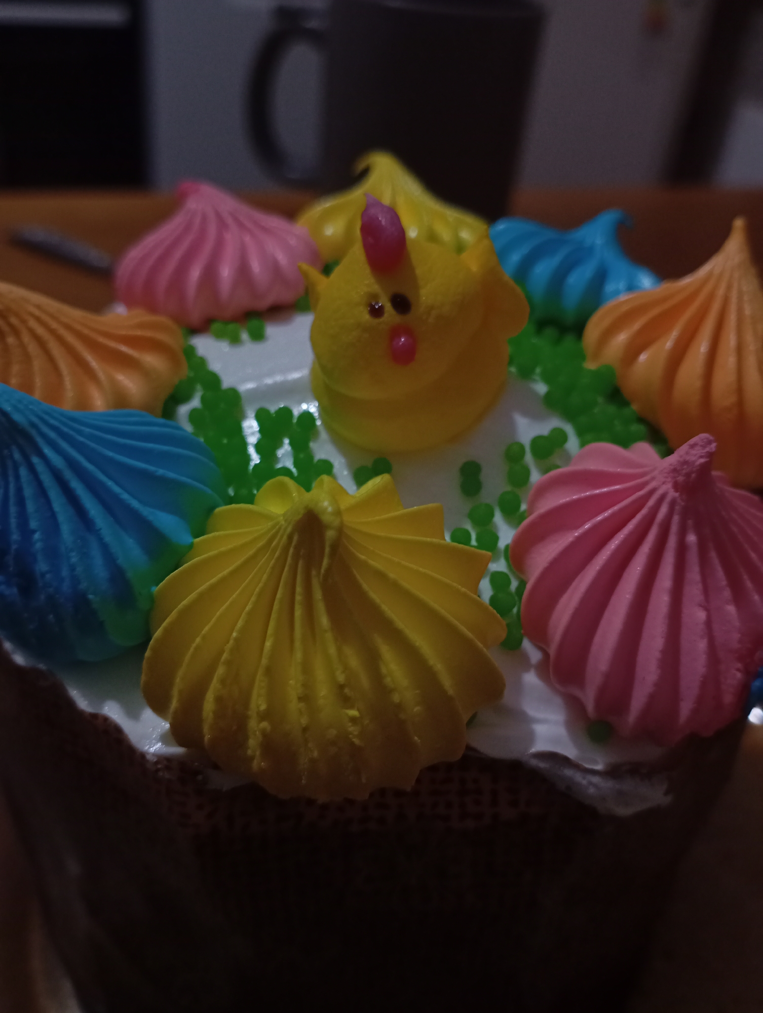 The image shows a close-up of an Easter cake decorated with colorful meringue swirls in shades of yellow, blue, pink, and orange. A yellow chick-shaped meringue sits in the center. Green sprinkles are scattered around the base of the decorations. The cake has white icing on top and a darker, textured base. A mug is visible in the blurred background. The location is unknown, though the user suggested the city of Krasnodar, Russia.