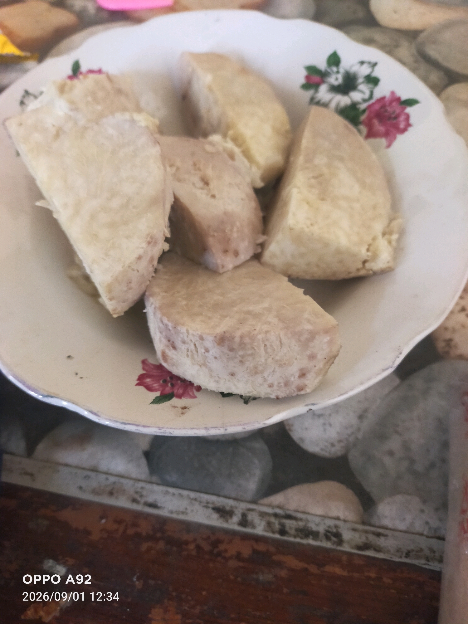 A close-up, overhead shot captures a white, scalloped-edge plate containing several pieces of boiled taro root, cut into wedges and cylindrical shapes. The taro is light tan in color with a slightly textured surface. The plate is decorated with a small floral pattern featuring pink and white flowers with green leaves. The plate rests on a surface that appears to be composed of smooth, grey and white stones, with some appearing round and others flattened. In the background, parts of other objects are visible, including what looks like a pink item and a yellow item, blurred out of focus. The lower portion of the image reveals a wooden surface with a textured, dark brown grain. At the bottom left, overlaid text displays "OPPO A92 2026/09/01 12:34", indicating the phone model and date/time of the photograph. The scene is indoors, likely on a table or countertop, with the lighting suggesting a natural light source from above. No people or specific actions are depicted. The location in Binangun, Indonesia, cannot be determined from the visual information presented in the image.