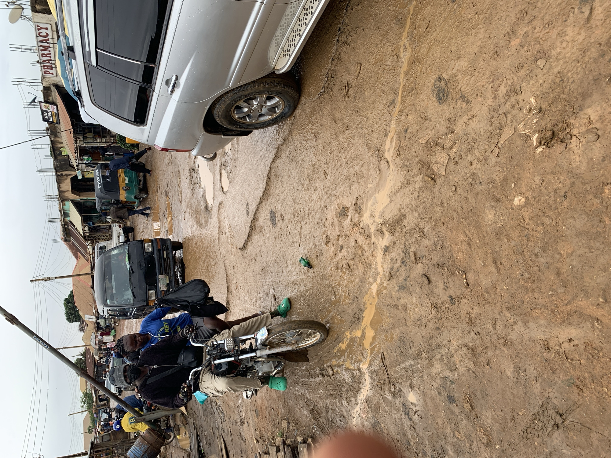 The image depicts a street in Rafin Par, Nigeria, characterized by muddy conditions. A silver SUV is partially visible in the upper left, parked on the side of the road. A black van is parked behind it. A motorcycle with two passengers is in the foreground. Both are wearing casual clothing; one in a blue sweatshirt, the other in a black jacket. Both wear green Crocs. A green bottle is on the ground near the motorcycle. Buildings with commercial signs, including one for a pharmacy, line the street. Other pedestrians and vehicles, including a yellow tricycle taxi, are also present.