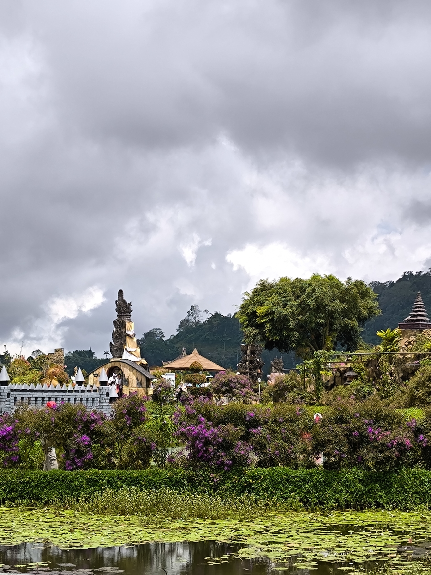 The image shows a water garden at Ulun Danu Beratan Temple in Bedugul, Indonesia. The temple complex is visible in the background with ornate spires and traditional Balinese architecture. There is a lake in the foreground covered with lily pads. Bushes with purple flowers are between the lake and the temple. The sky is cloudy and overcast. There appear to be people within the temple complex.
