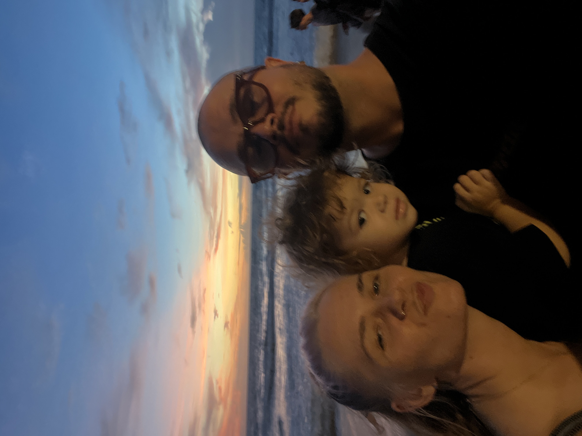 In this selfie, taken at twilight on a beach, a man, a woman, and a young child are featured prominently. The man, in the foreground on the right, is bald, has a beard, and wears glasses. The woman is on the left, and the child is held between them. In the background, the ocean is visible with waves crashing on the shore. The sky displays a beautiful sunset with hues of pink, orange, and blue, and scattered clouds. Other people are visible in the background on the beach.